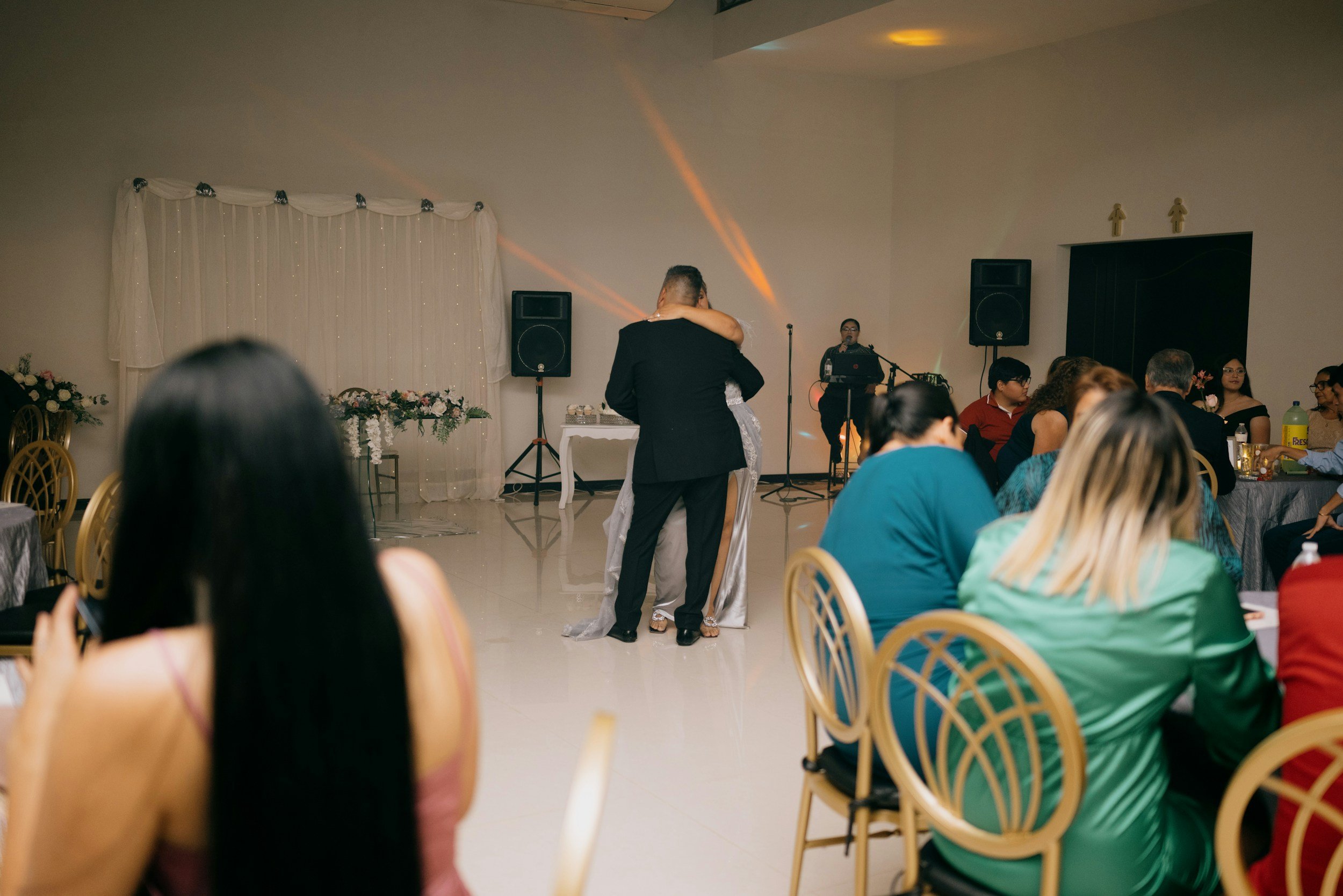 Couple dancing at a wedding reception with guests seated at tables in the background, decorated with flowers and elegant drapery, while a DJ plays music.