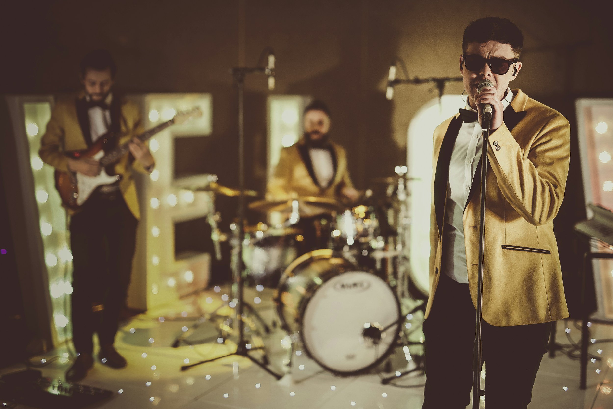 A band performing in a dimly lit indoor venue. The lead singer, wearing sunglasses and a gold tuxedo jacket, is singing into a microphone. In the background, a guitarist and drummer are visible.