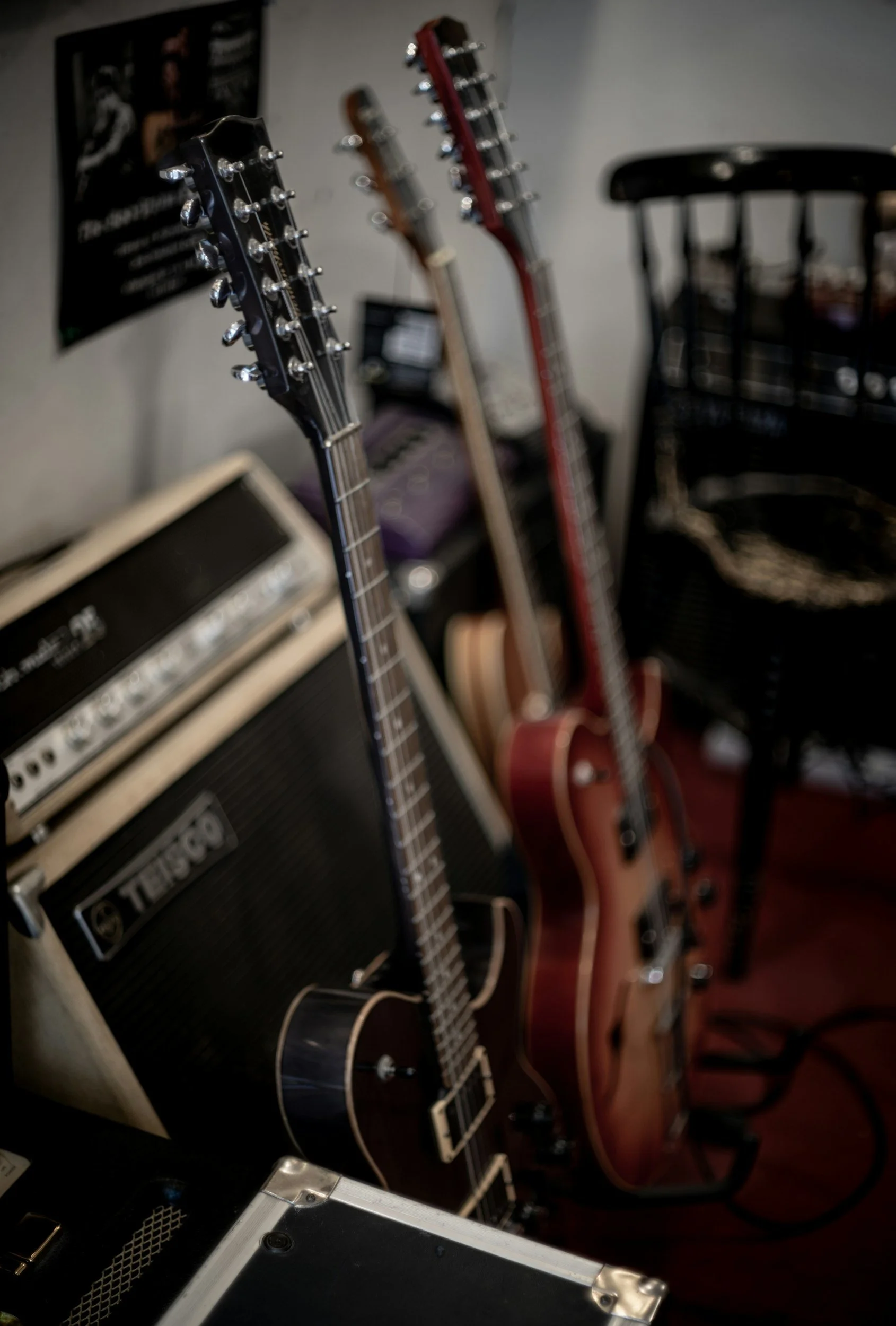 Close-up of guitar headstocks, including a black Epiphone guitar, with a guitar amplifier and a wooden chair in the background.