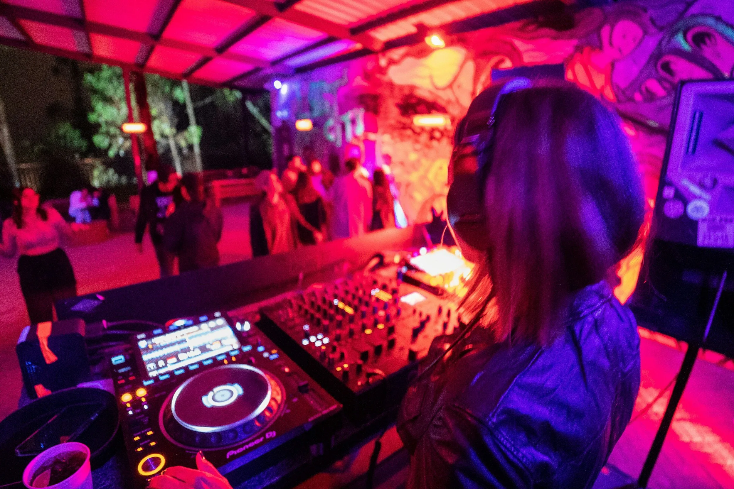 A DJ with long hair wearing headphones is mixing music at a party with colorful lighting. People are dancing and socializing in the background.