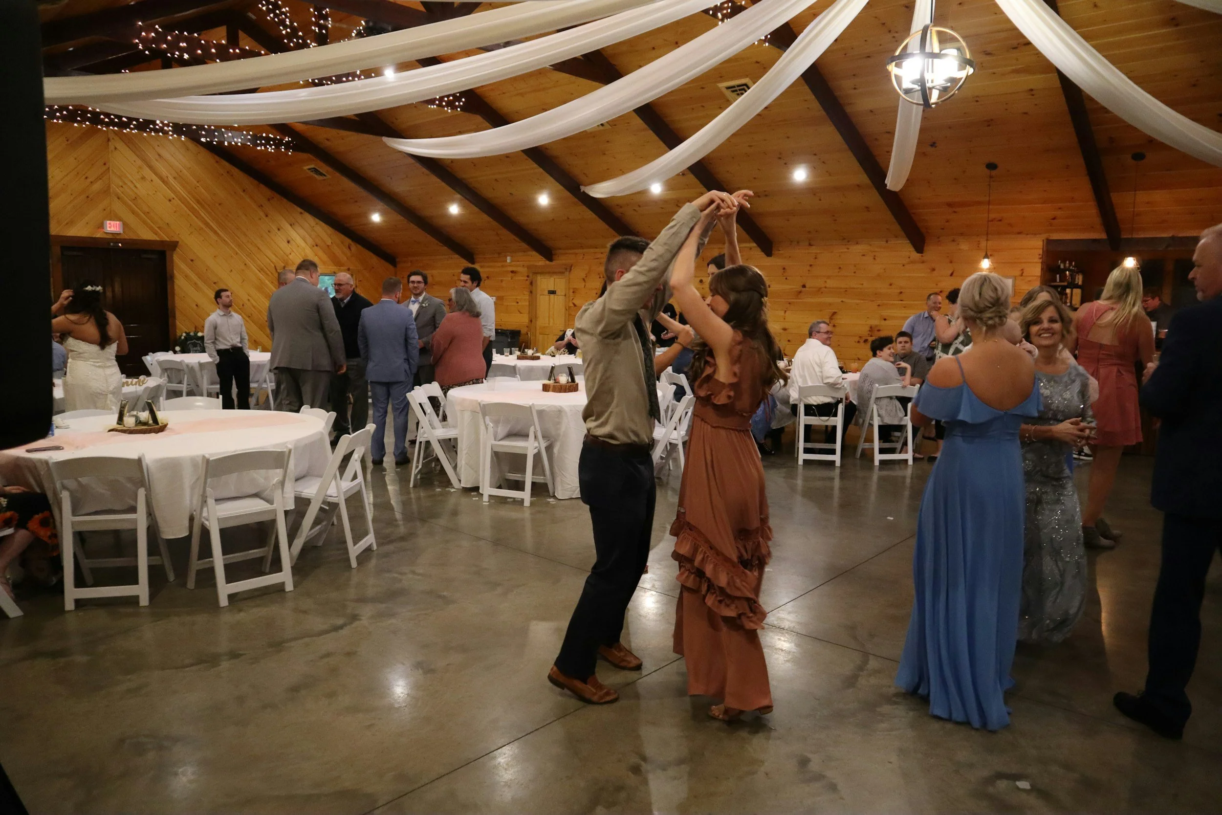 People dancing and socializing at a wedding reception in a barn with wooden walls and ceiling, with string lights and draped fabric decorations.