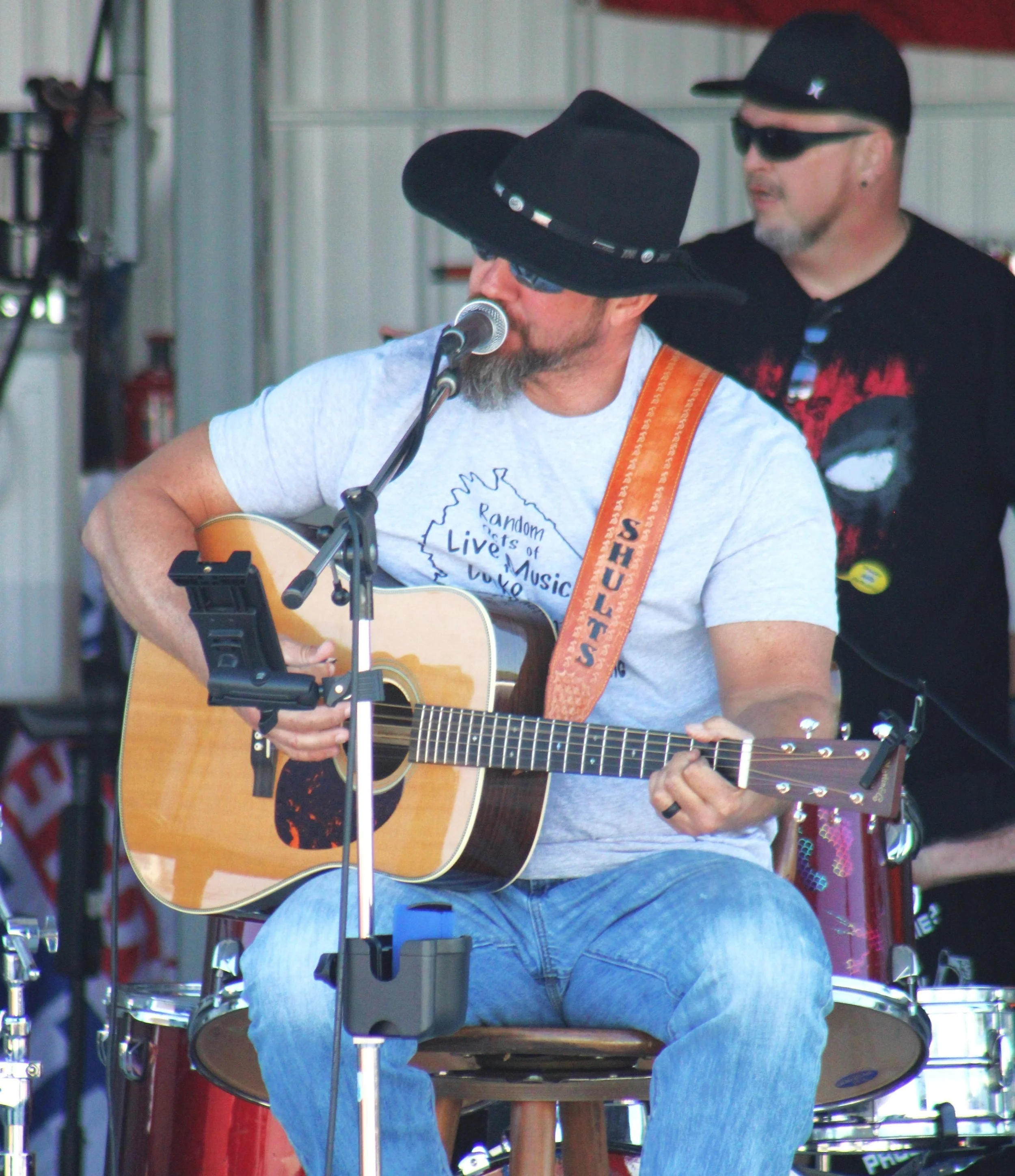 A man wearing a black wide-brimmed hat, sunglasses, and a light gray T-shirt with a graphic of a map and text, is playing an acoustic guitar and singing into a microphone. He is seated, and another man wearing a black cap, black sunglasses, and a black T-shirt stands behind him.