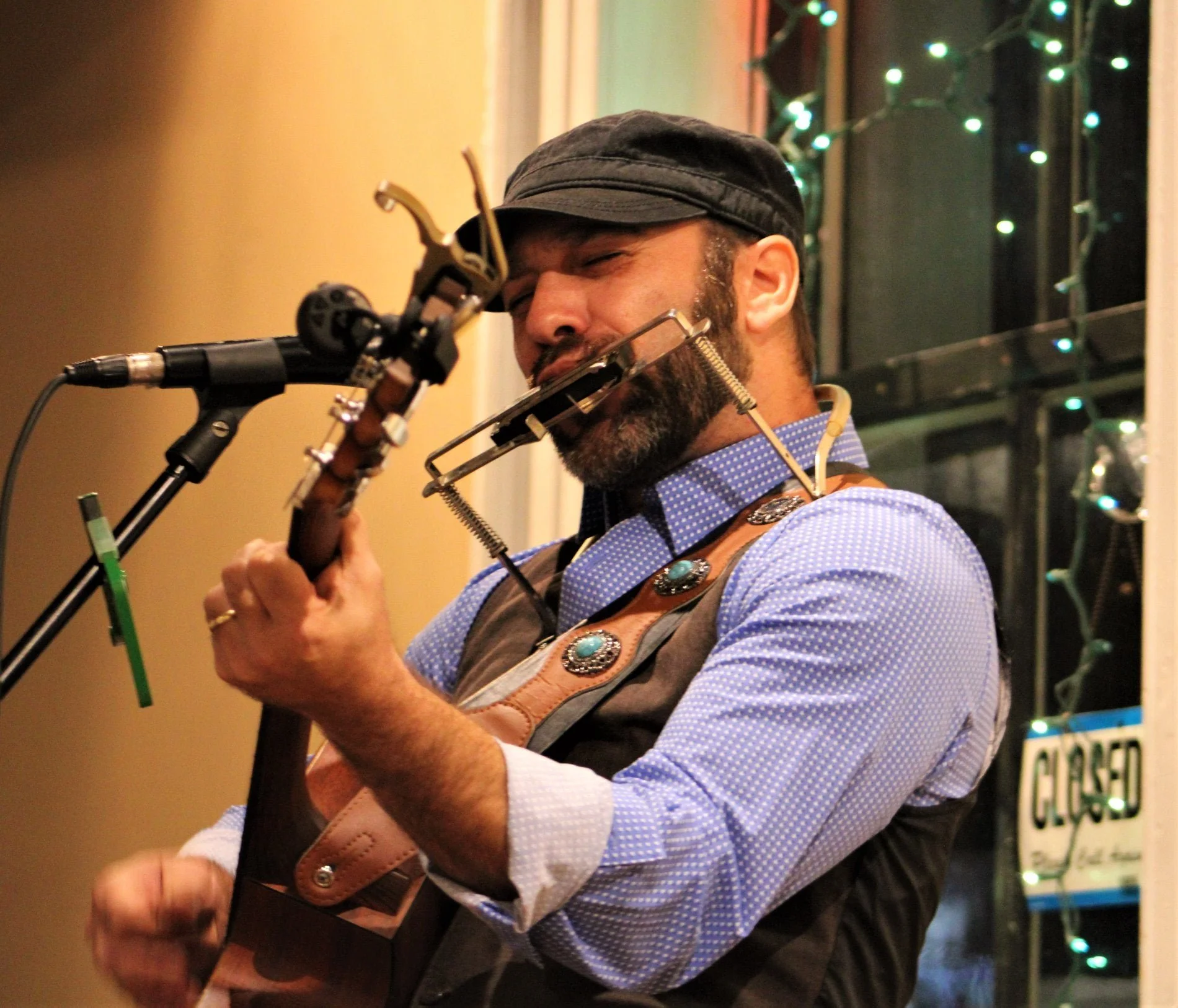 A man with a beard and mustache, wearing a black cap and a blue checkered shirt, playing a guitar with a harmonica holder around his neck. He is singing into a microphone, with colorful string lights and a sign with partially visible text in the background.