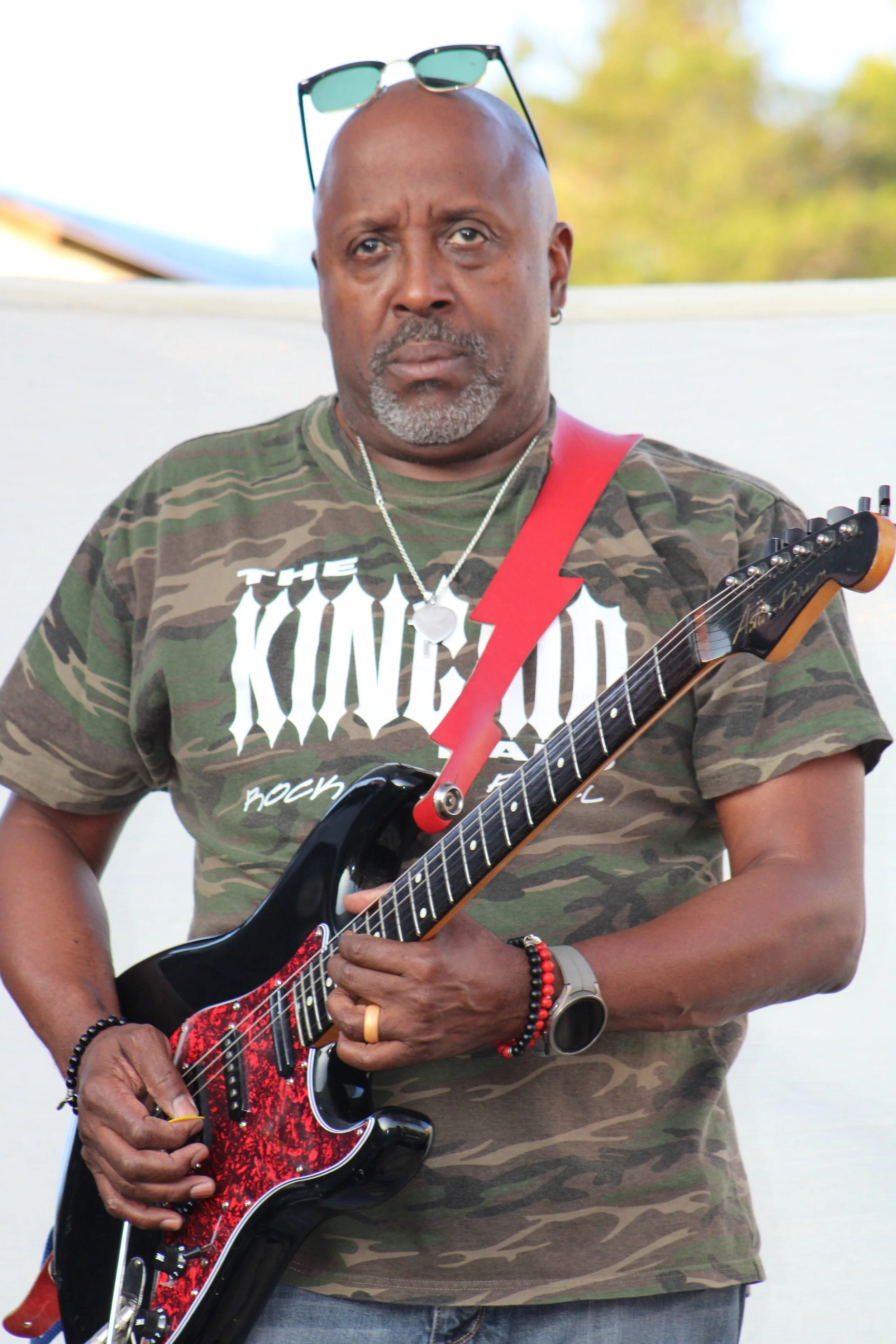 A man with a bald head, gray beard, wearing sunglasses on his head, a camouflage T-shirt, and various jewelry, playing an electric guitar outdoors with a blurred background of trees and sky.