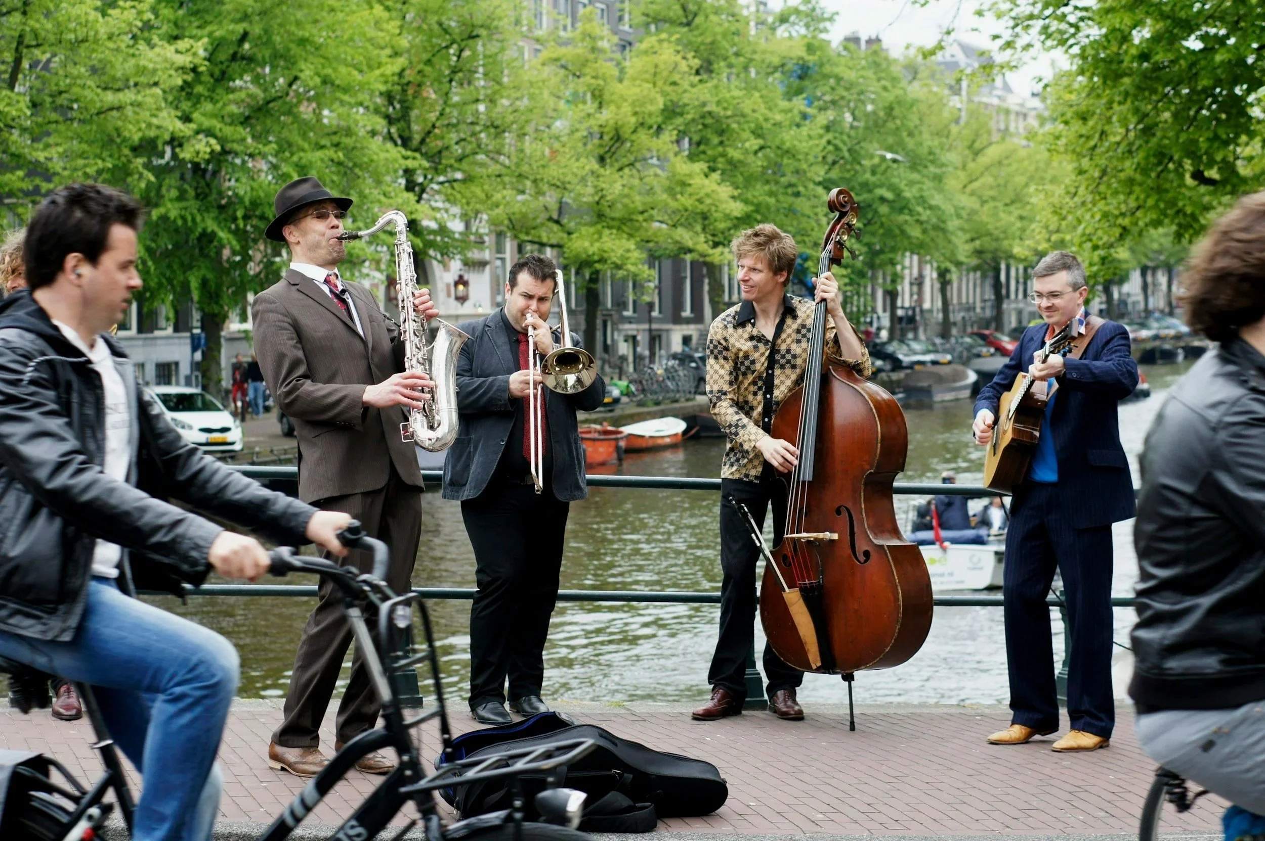 A group of street musicians performing near a canal, with two men playing saxophone and double bass, and others playing guitar and trumpet, in a city with green trees and buildings in the background.