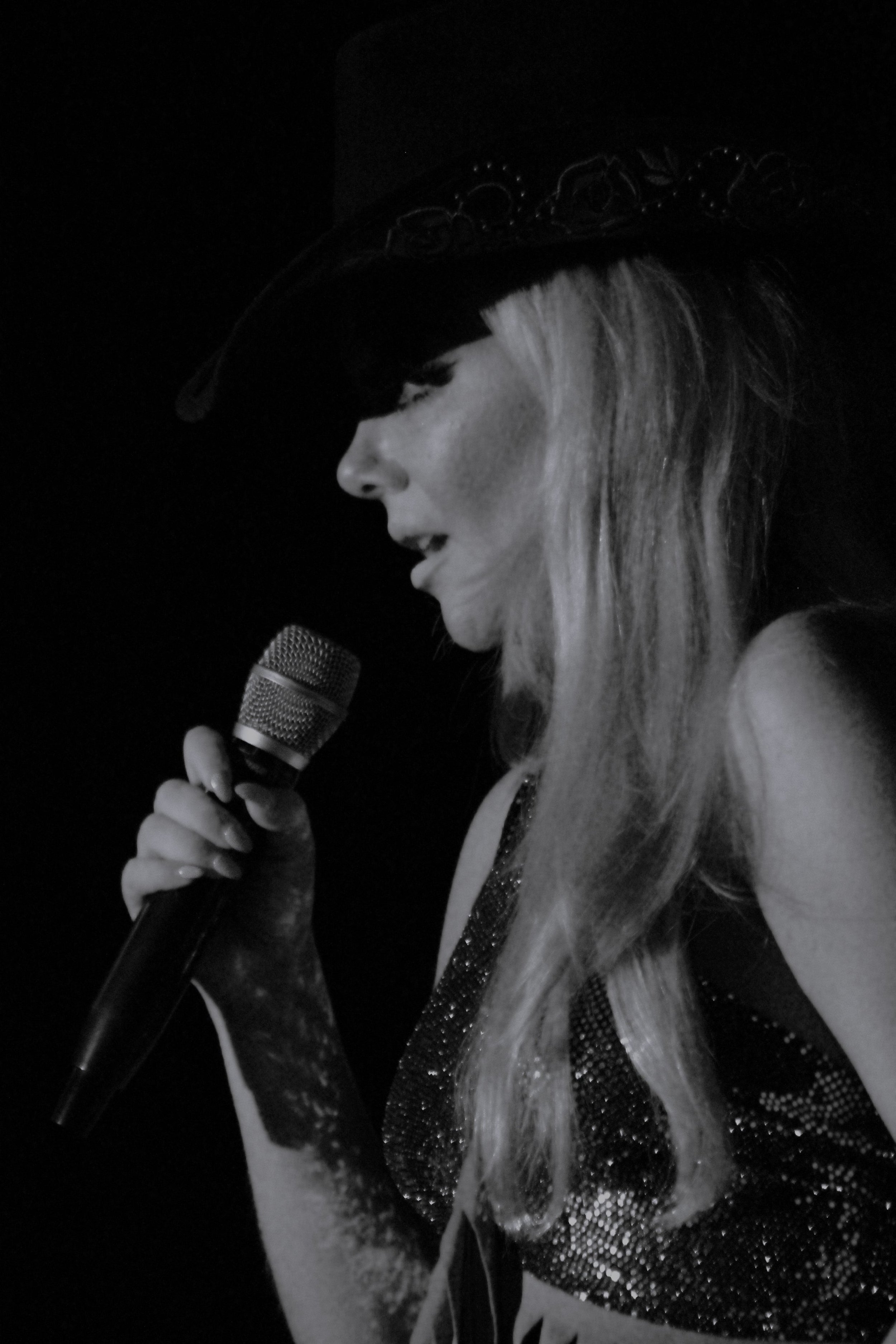 A woman with long blonde hair wearing a decorative hat and a shiny, sparkly top, singing into a microphone.