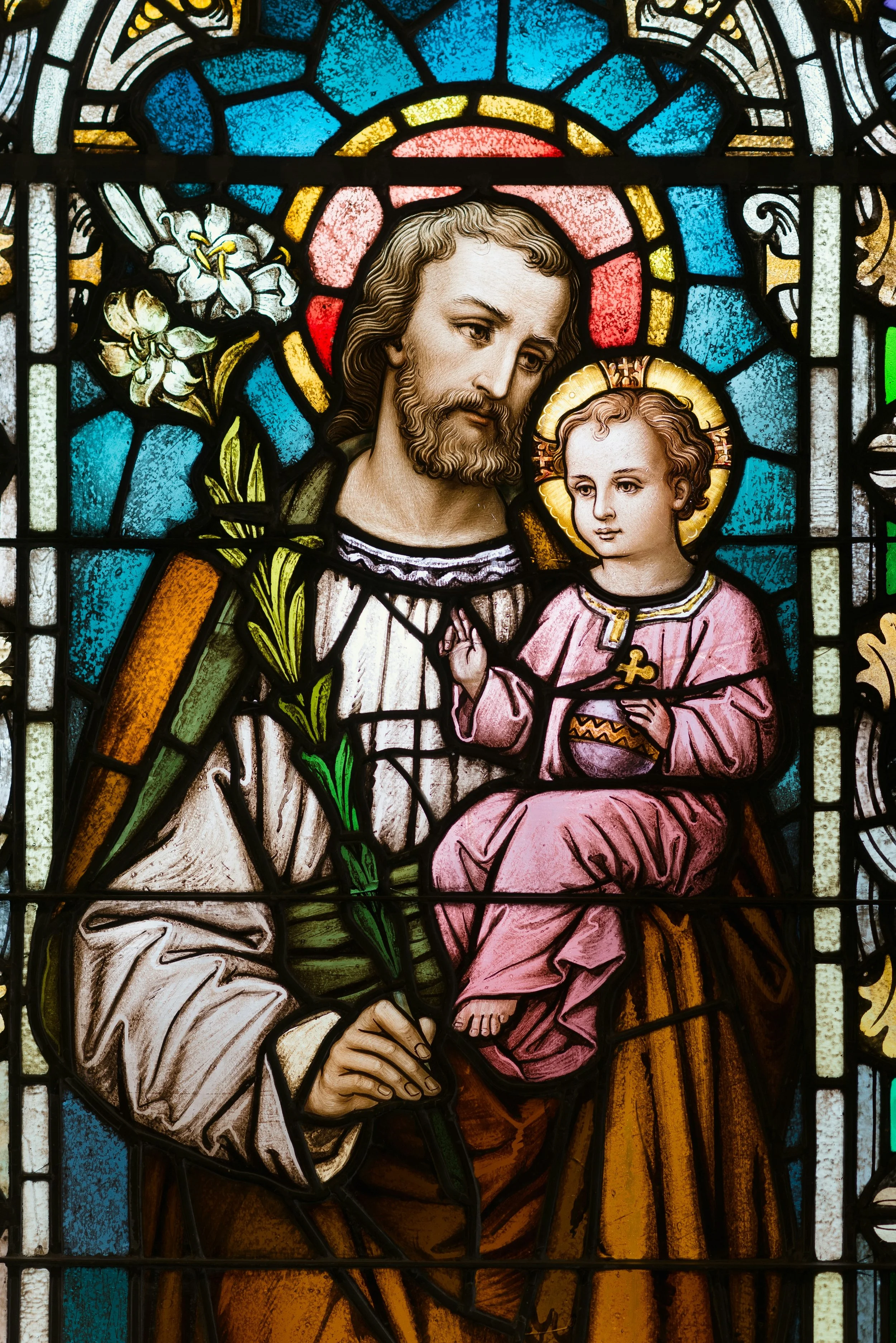 Colorful stained glass window depicting a religious scene with a bearded man holding a lily and a young boy with a halo, both dressed in robes.