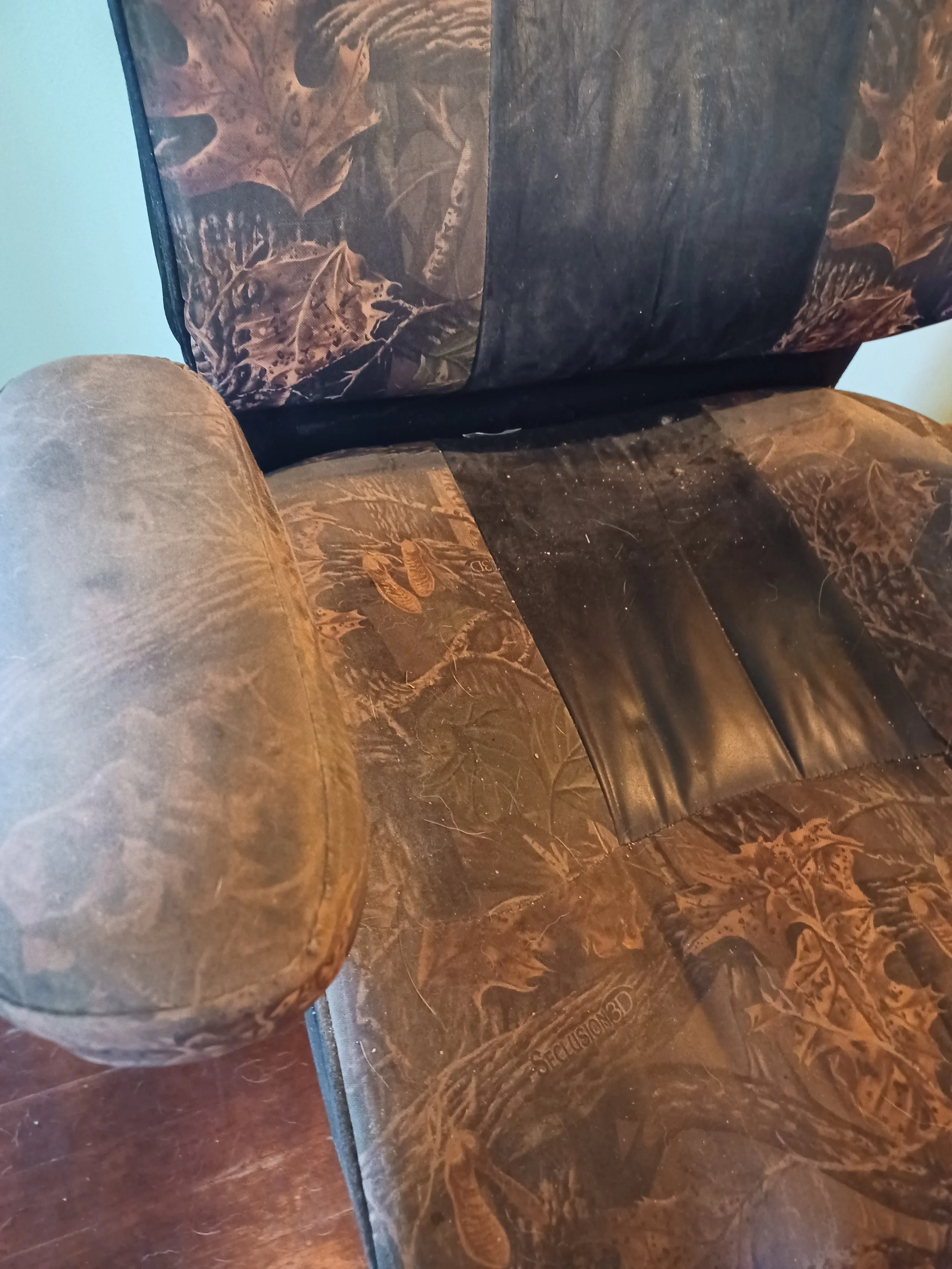 Close-up photo of a camouflage-patterned chair with a cylindrical armrest and a section of the seat visible, placed on a wooden floor.  Visibly dirty.