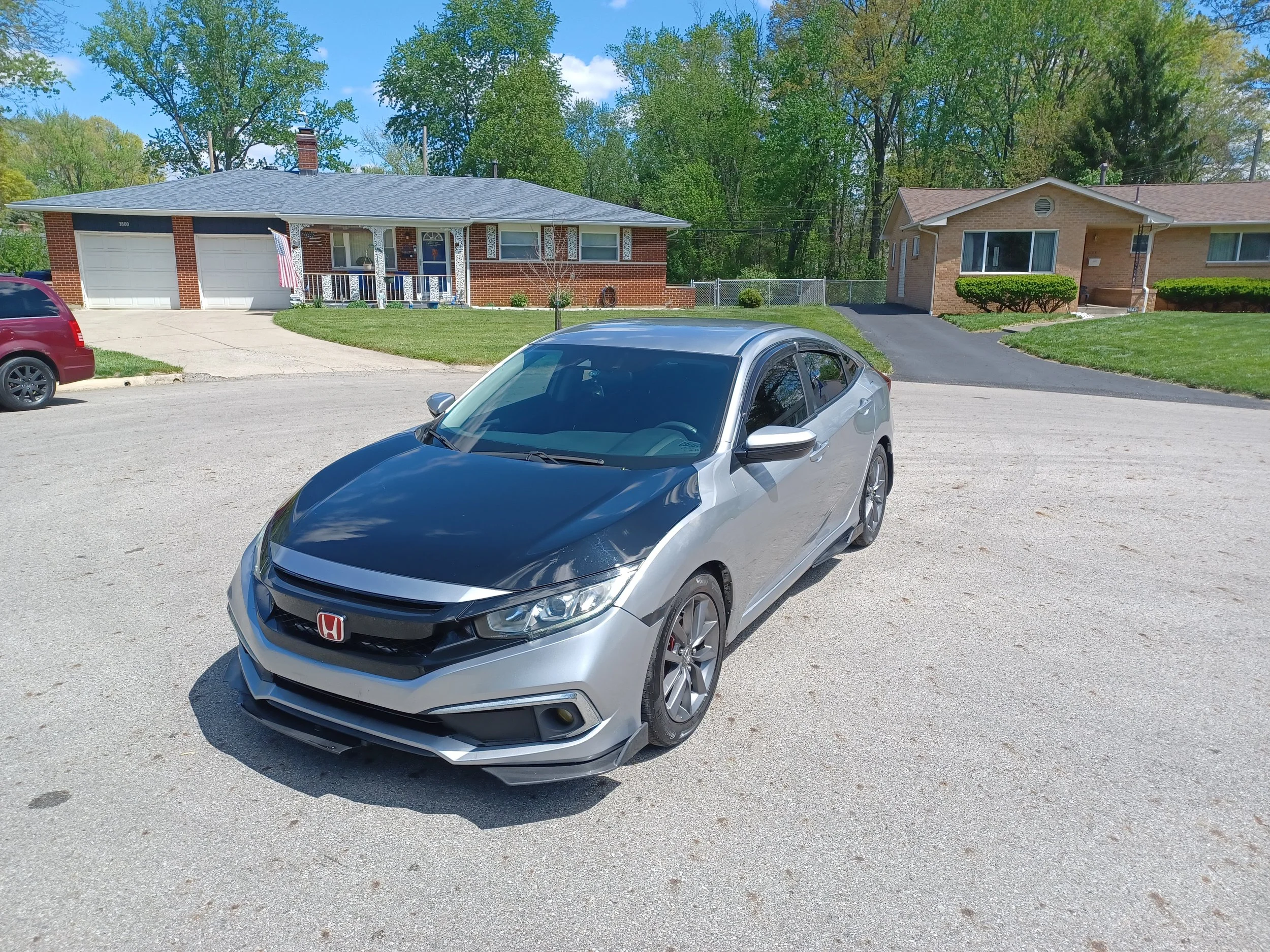 A silver Honda Civic parked in a gravel lot in front of two suburban houses with green trees in the background.