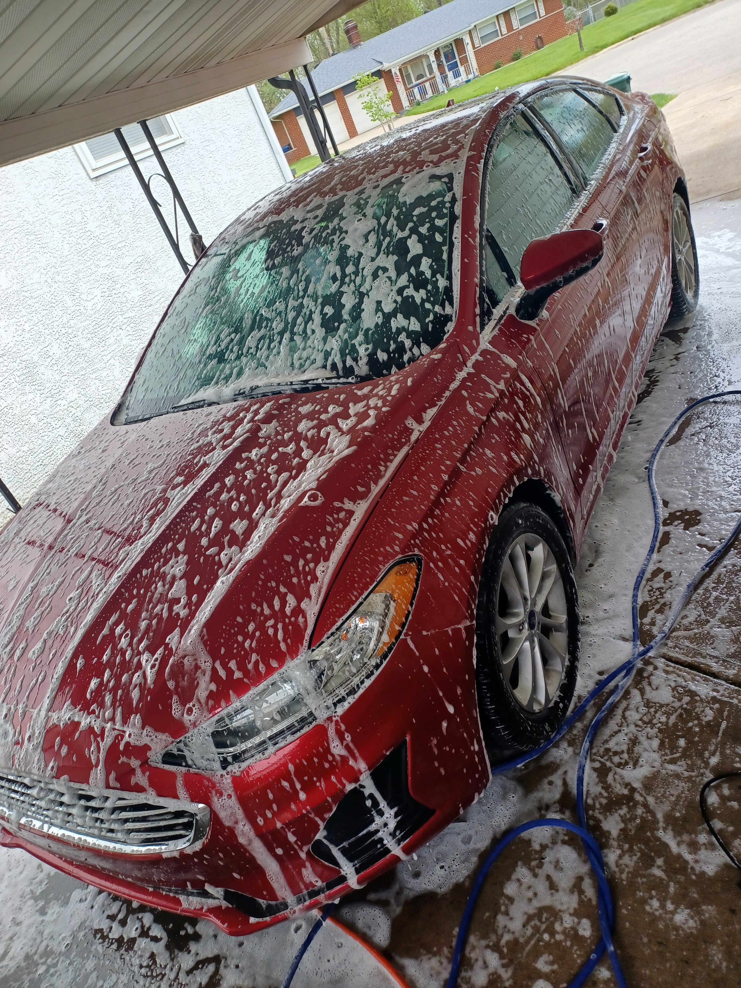 Red car being washed outside with soap and water, surrounded by hoses and suds, in a residential driveway.