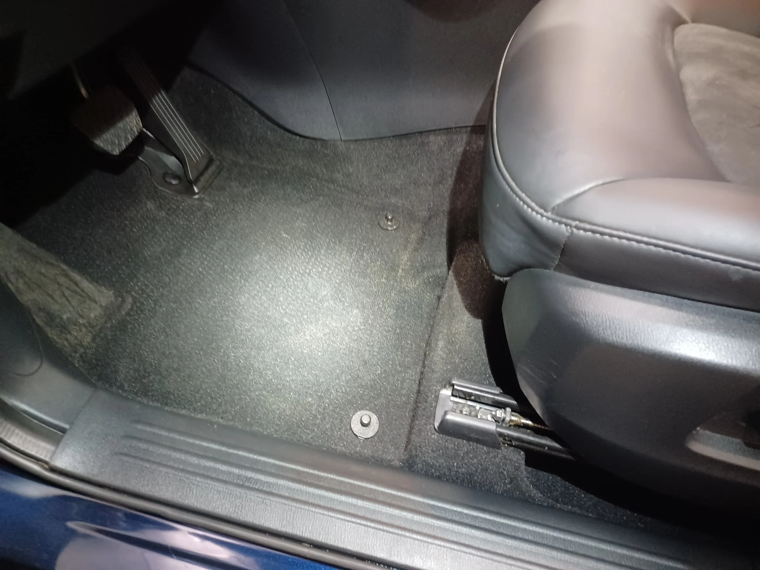 The image shows the interior of a car focusing on the passenger side footwell area. There is a black floor mat, part of a gray seat on the right, a vehicle's pedal on the left, and some metal fasteners on the floor. A black wire runs across the floor