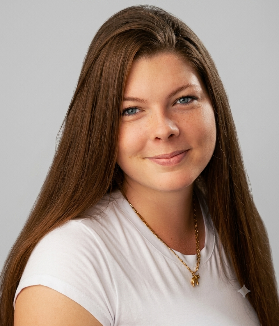 Portrait of a young woman with long brown hair, light blue eyes, and freckles, wearing a white T-shirt and a gold necklace with a small pendant, smiling softly against a plain gray background.