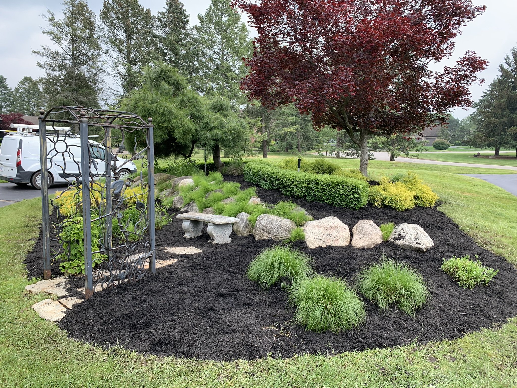 A landscaped garden with a small flowerbed bordered by large rocks, various green plants, a bush, and three trees, one with red leaves, in a park setting with a pathway, grass, and a street with parked cars and houses in the background.