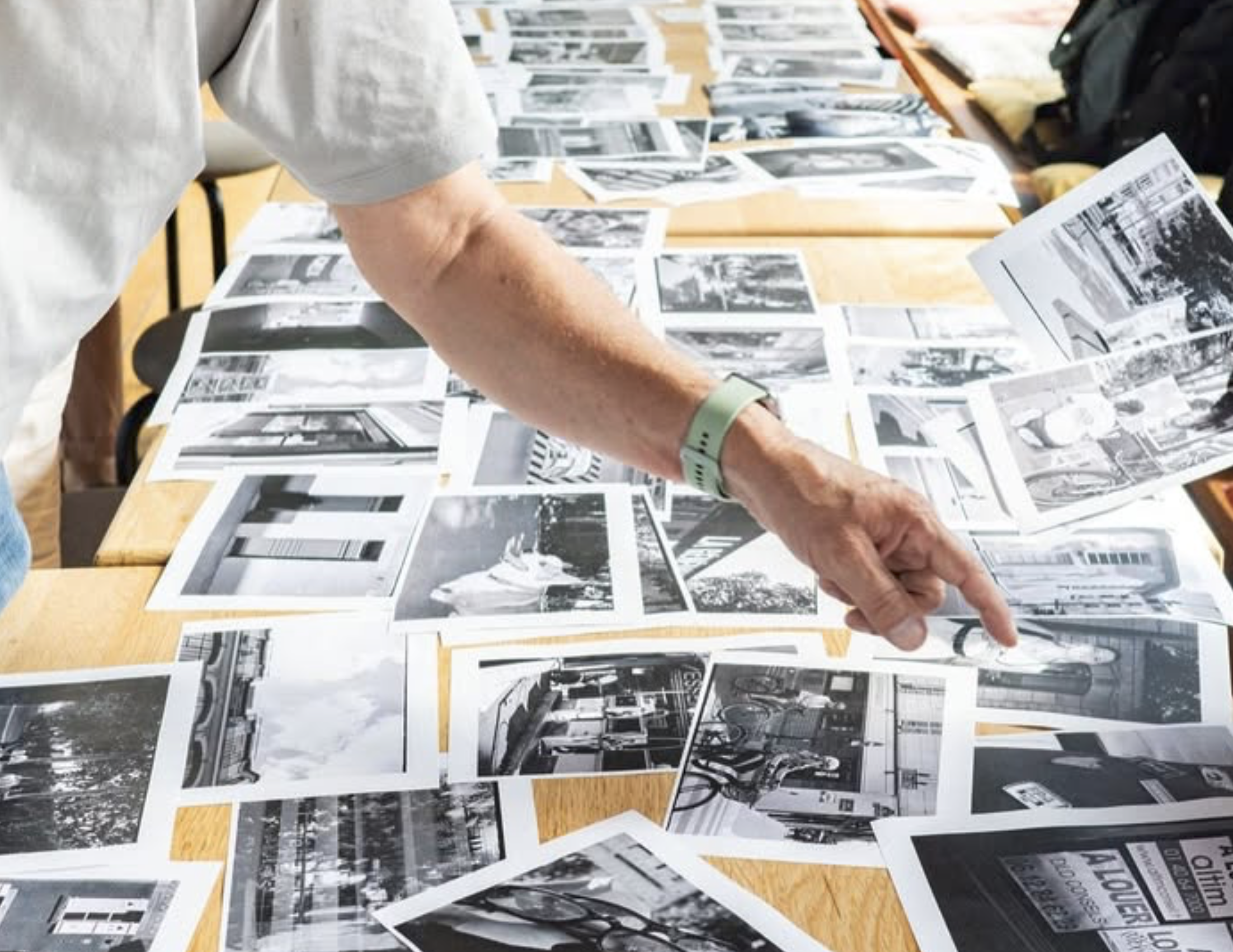 Une personne consulte un affichage de photographies en noir et blanc sur une table en bois clair. La personne porte un bracelet vert et une chemise claire à manches longues. Des sacs et autres objets sont visibles à l'arrière.