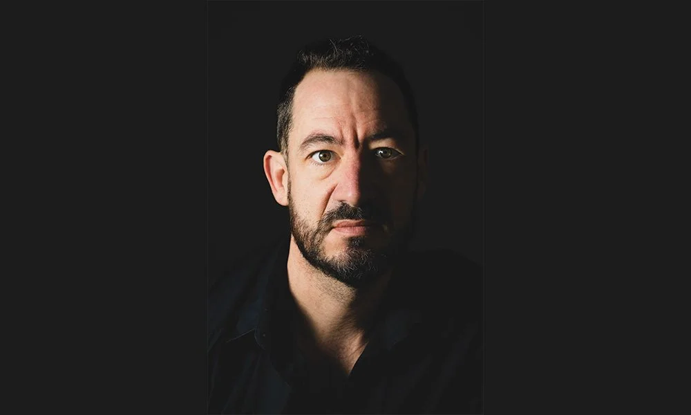 Portrait d'un homme avec une barbe et une expression neutre, en noir et blanc, contre un fond noir