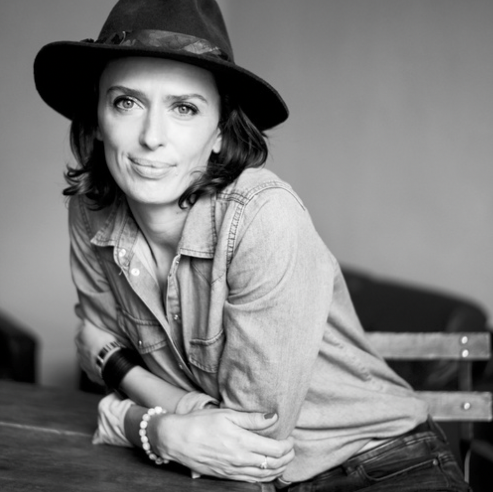Une femme portant un chapeau noir, une veste en denim, un bracelet brillant et une montre, assise sur une surface, regardant l'objectif dans une photo en noir et blanc.