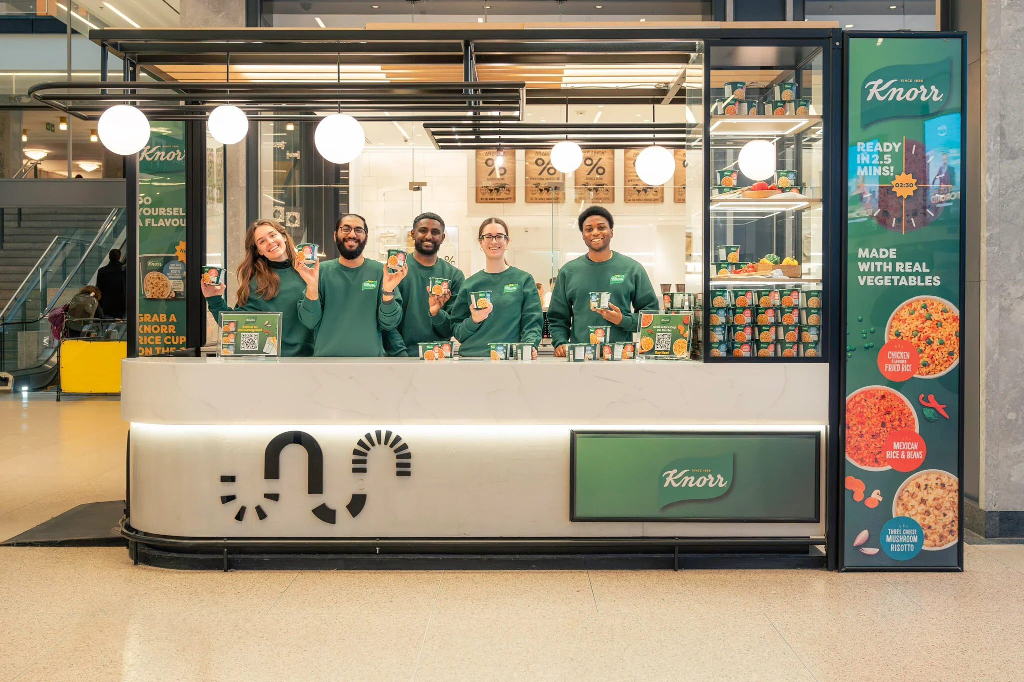 A group of five smiling employees in green uniforms working at a Knorr food stand at an indoor location, holding small containers of a food product, with various promotional signs and food images displayed.