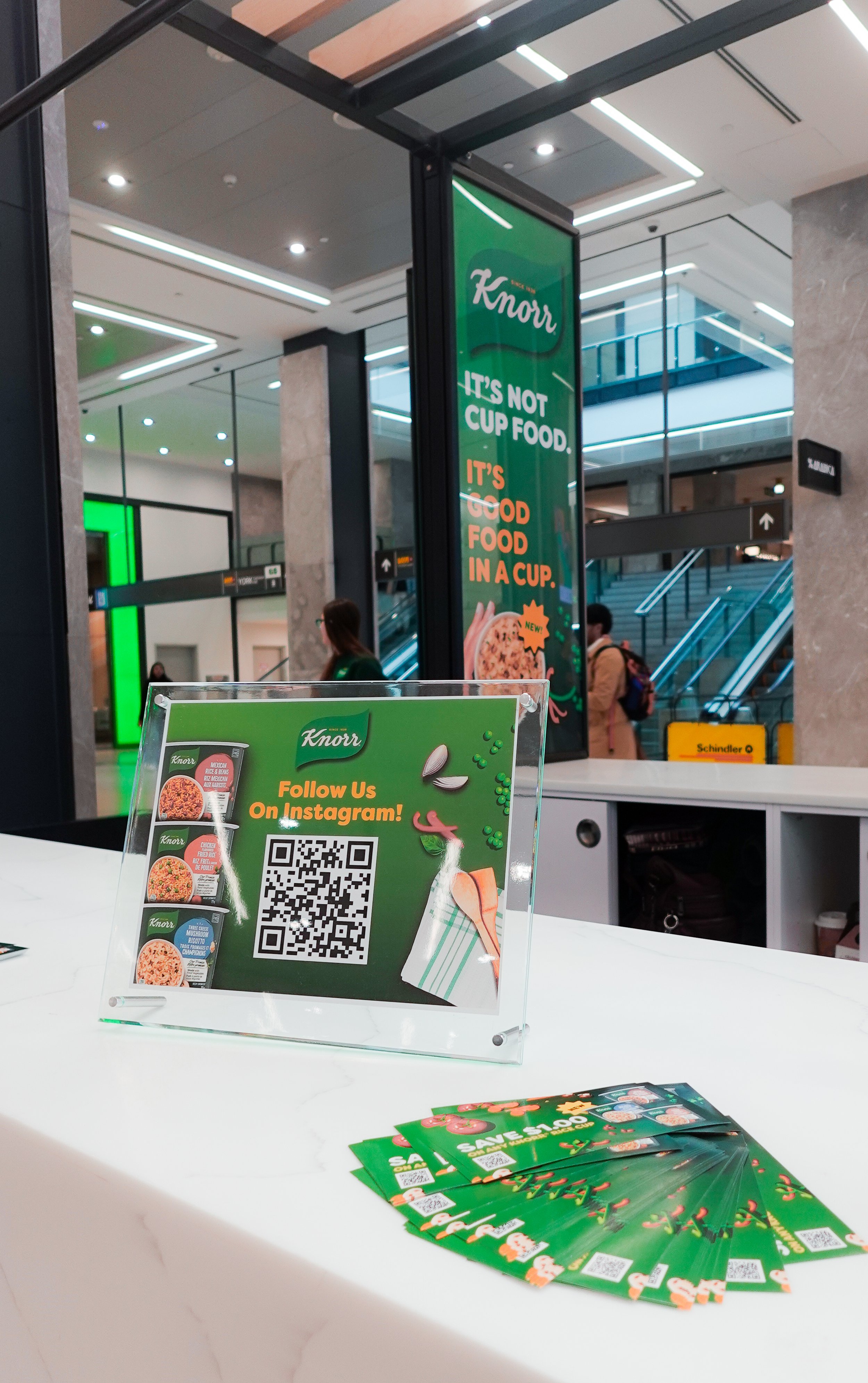 A promotional display on a counter for Knorr, featuring a QR code and promotional flyers with a green background. The flyers promote discounts and include graphics of food ingredients like vegetables and a drink, designed by Sunnii Studios.
