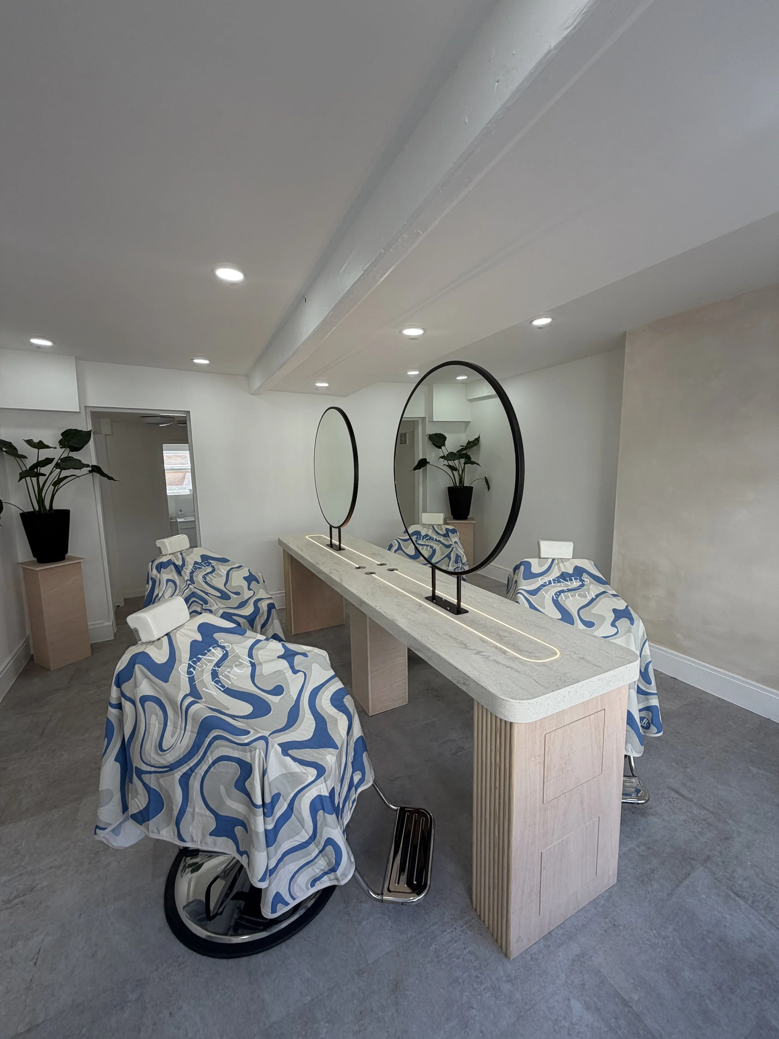 A room with a beige countertop island that has wave-patterned cloths covering chairs around it, two circular mirrors with black frames, a potted plant on a side table, and ceiling lights.