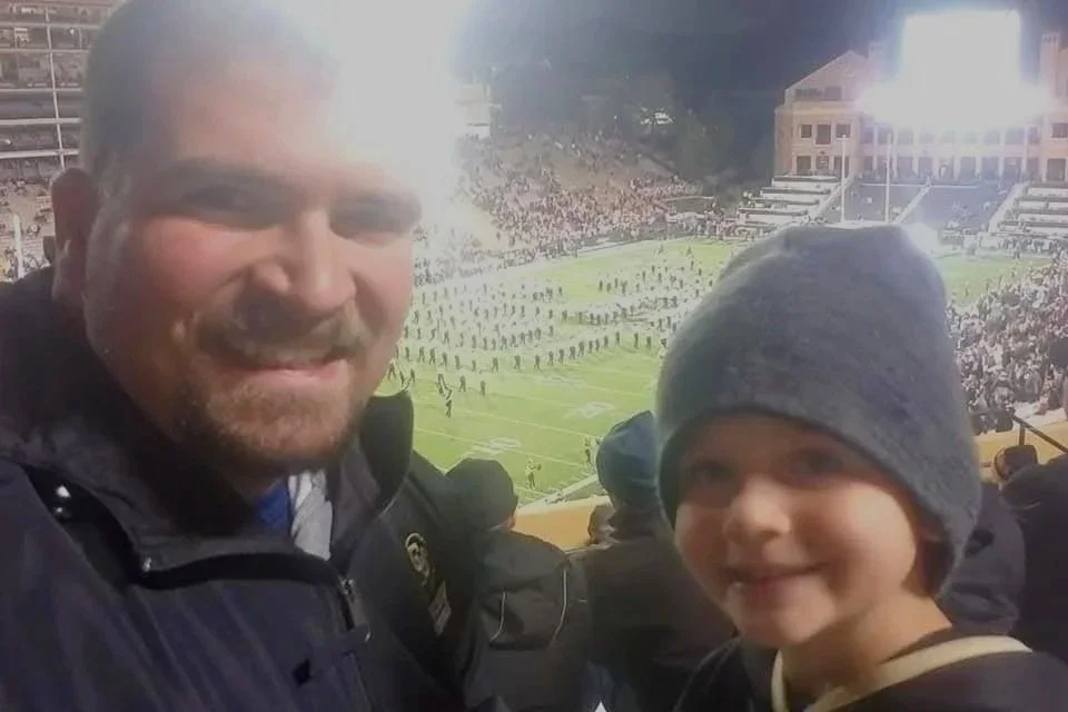 A man and a young boy smiling at a football game in a stadium. The man has a beard and is wearing a dark jacket, and the boy is wearing a gray beanie. The stadium is filled with spectators and a football field is visible in the background.