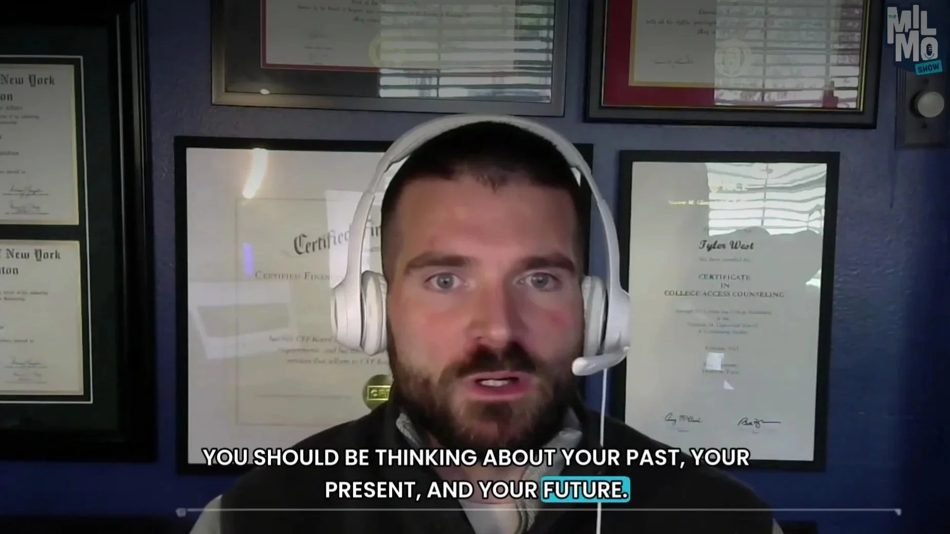 A man wearing headphones with a microphone, speaking during a virtual meeting, with a wall of framed certificates behind him.