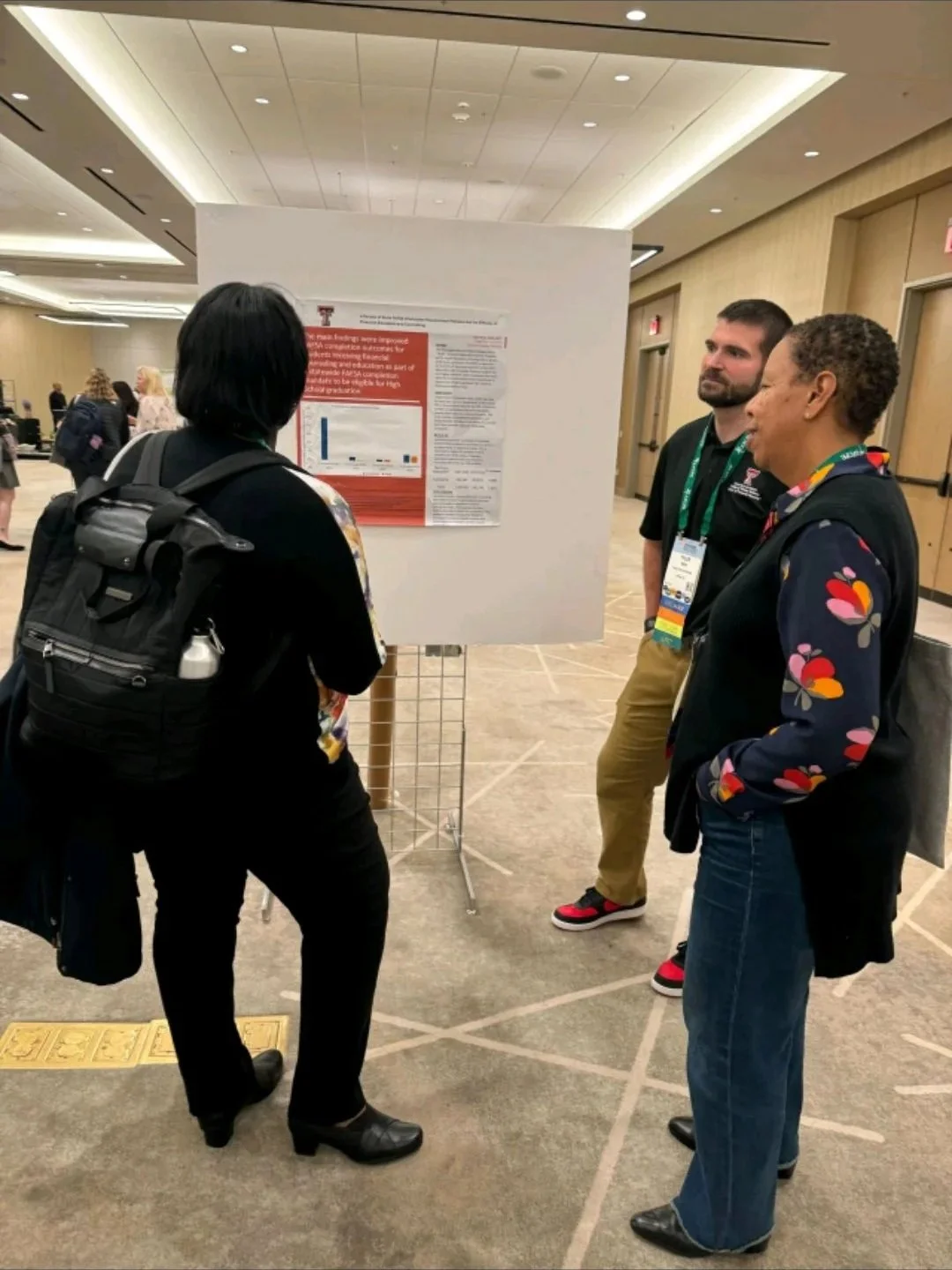 Three people standing in front of a research poster at a conference in a large well-lit room with a high ceiling. One person with a backpack is speaking to two others, one of whom has a colorful jacket and the other has a lanyard with a name badge.