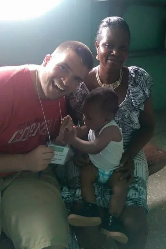 A Caucasian man in a red shirt and a woman with dark hair, wearing a patterned dress, sitting together with a young child, possibly of African descent, who is playing with the man's hand and appears to be holding a card or ID. The setting looks like an indoor or semi-outdoor space with green walls and sunlight coming in.