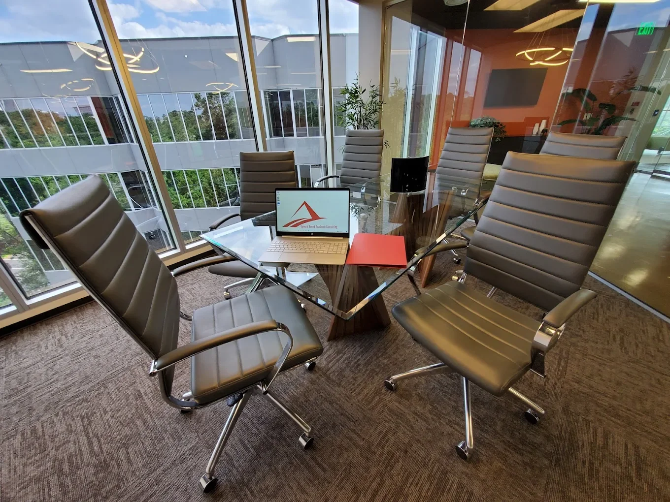 Modern glass conference room with six black leather office chairs around a glass table, a laptop, a red folder, a black speaker, and a water bottle. Floor-to-ceiling windows show a building and trees outside.