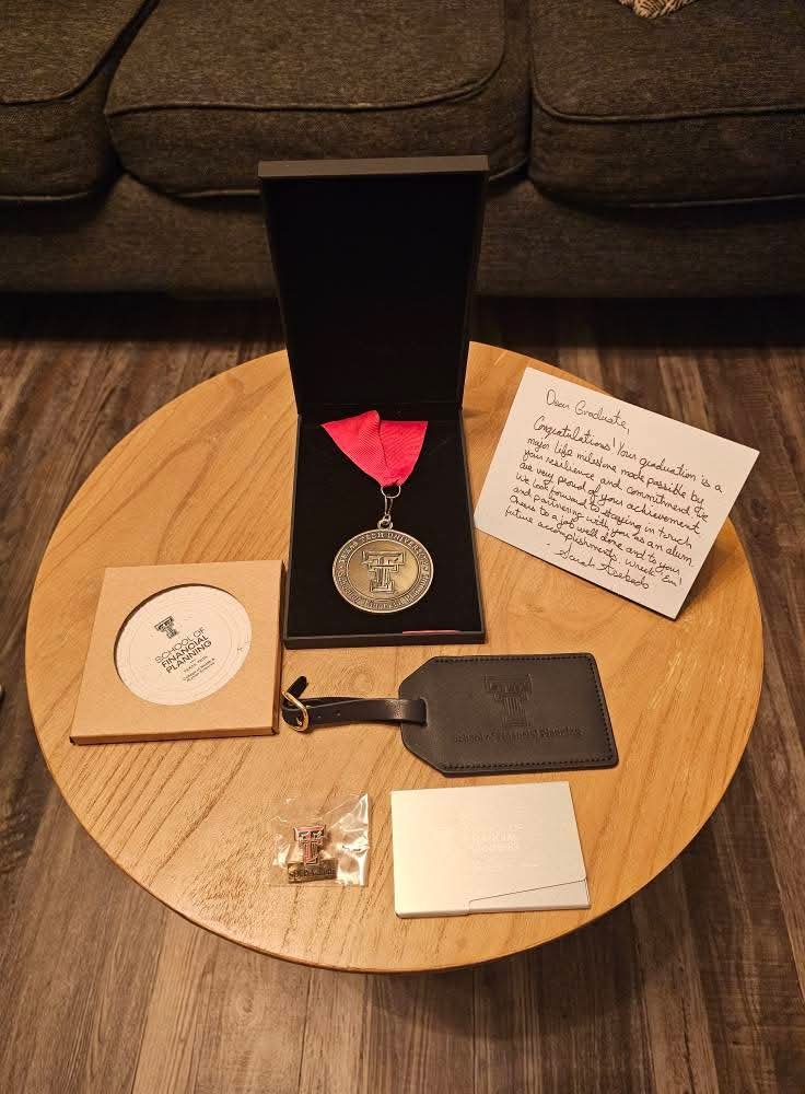 A Texas Tech University medal with a red ribbon in a black box, a handwritten note congratulating an graduate, a black luggage tag, a small pin, and several small cards on a wooden table.