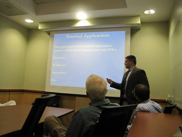 A man in a suit giving a presentation on practical applications of principles, with a slide projected on the wall behind him, in a conference room with several seated attendees.