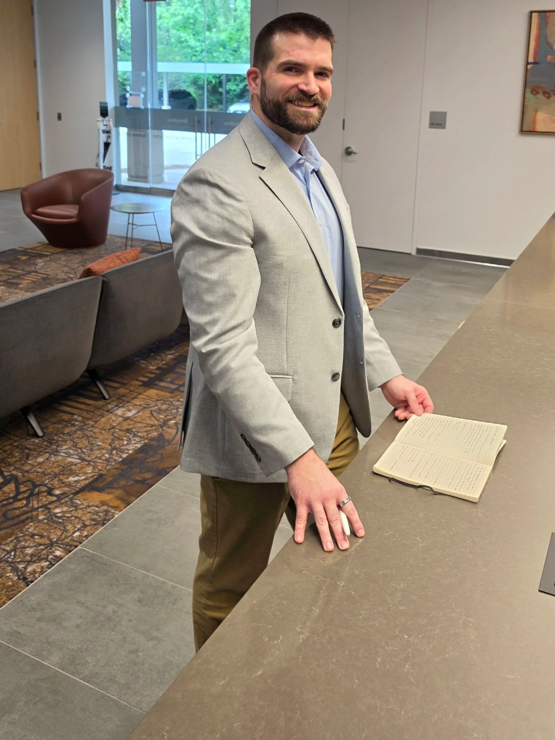 A man with a beard in a gray blazer standing in a modern office lobby, resting his hand on a counter, with an open notebook in front of him, and chairs and a glass door behind him.