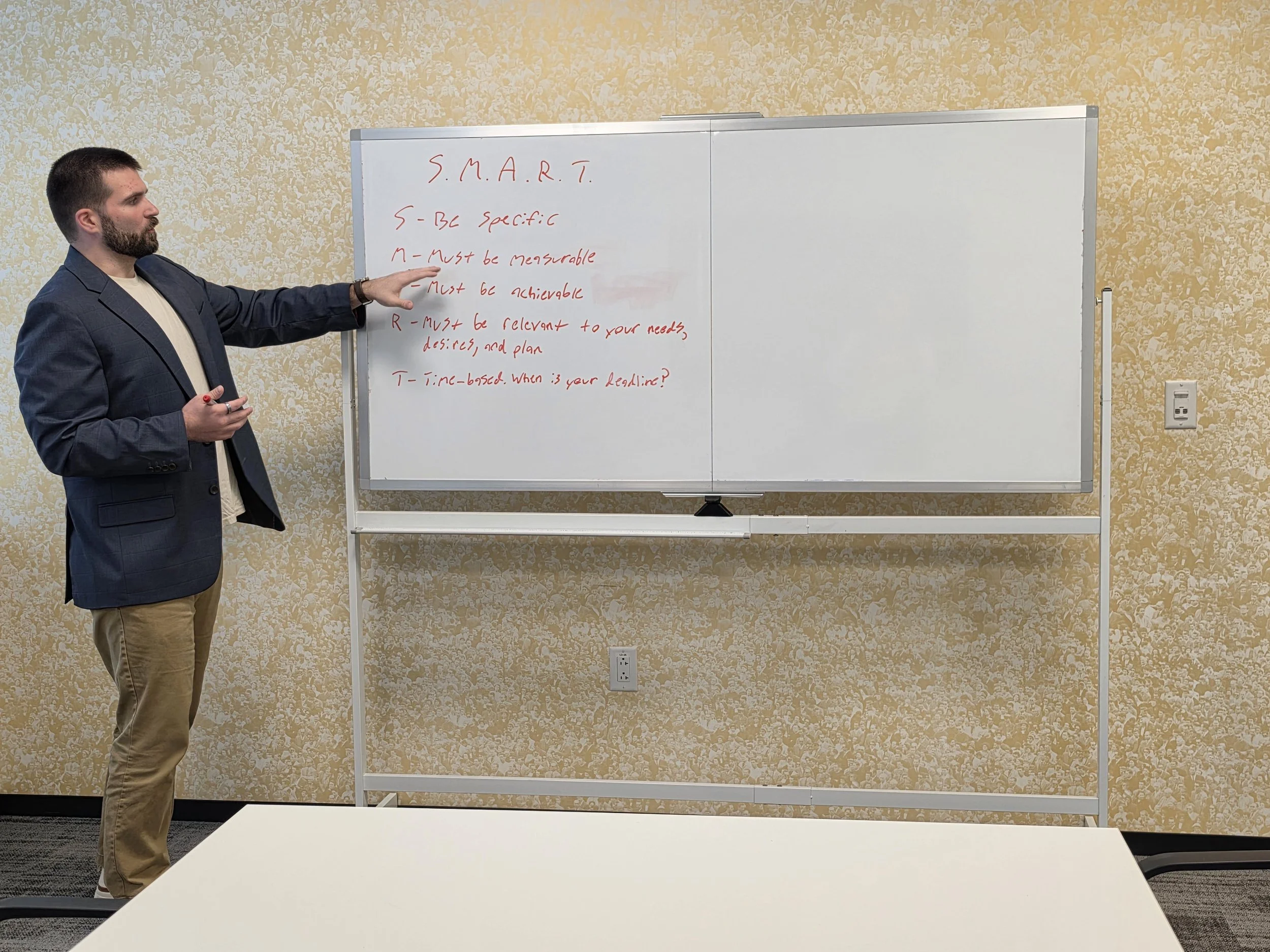 A man in a dark blazer and beige pants standing next to a whiteboard with red handwritten text about S.M.A.R.T. goals, pointing at the whiteboard in a room with beige patterned walls.