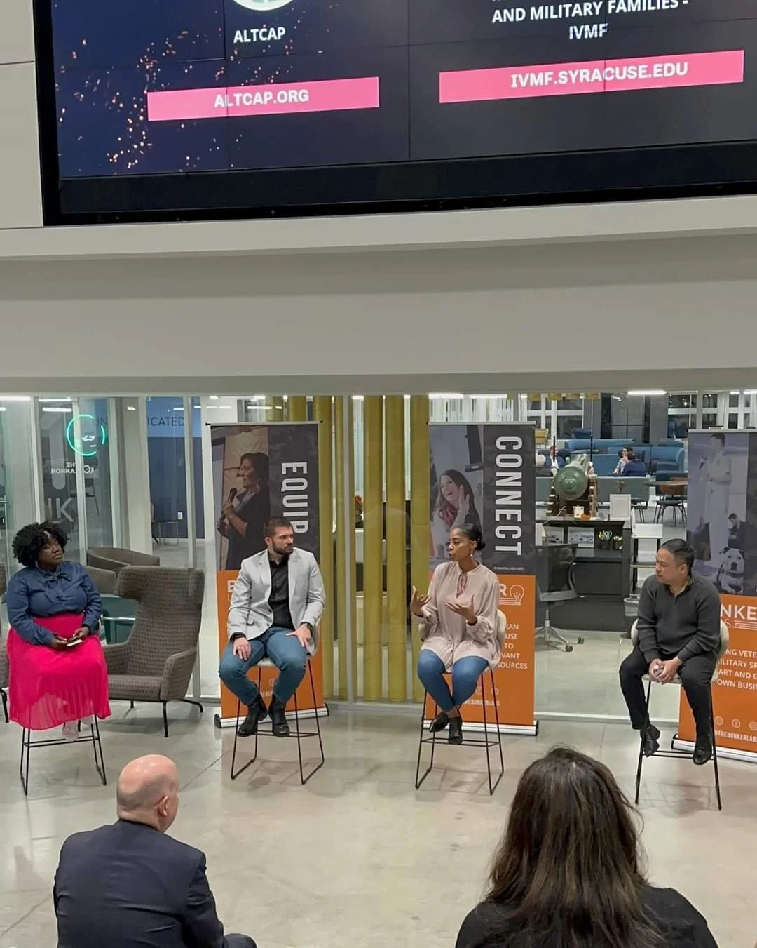 A panel discussion taking place indoors with four people sitting on chairs on stage. There are two large vertical banners behind them with images of women. An audience is seated in front, with two people visible from behind. The background shows a mo