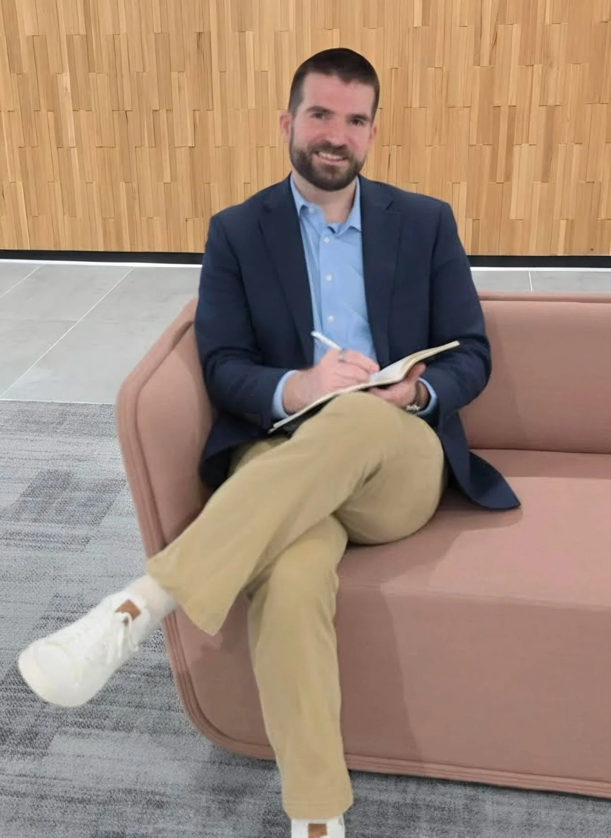 A man with dark hair and beard, dressed in a blue blazer and khaki pants, sitting on a pink couch, holding a notebook and pen, with a wooden wall background.