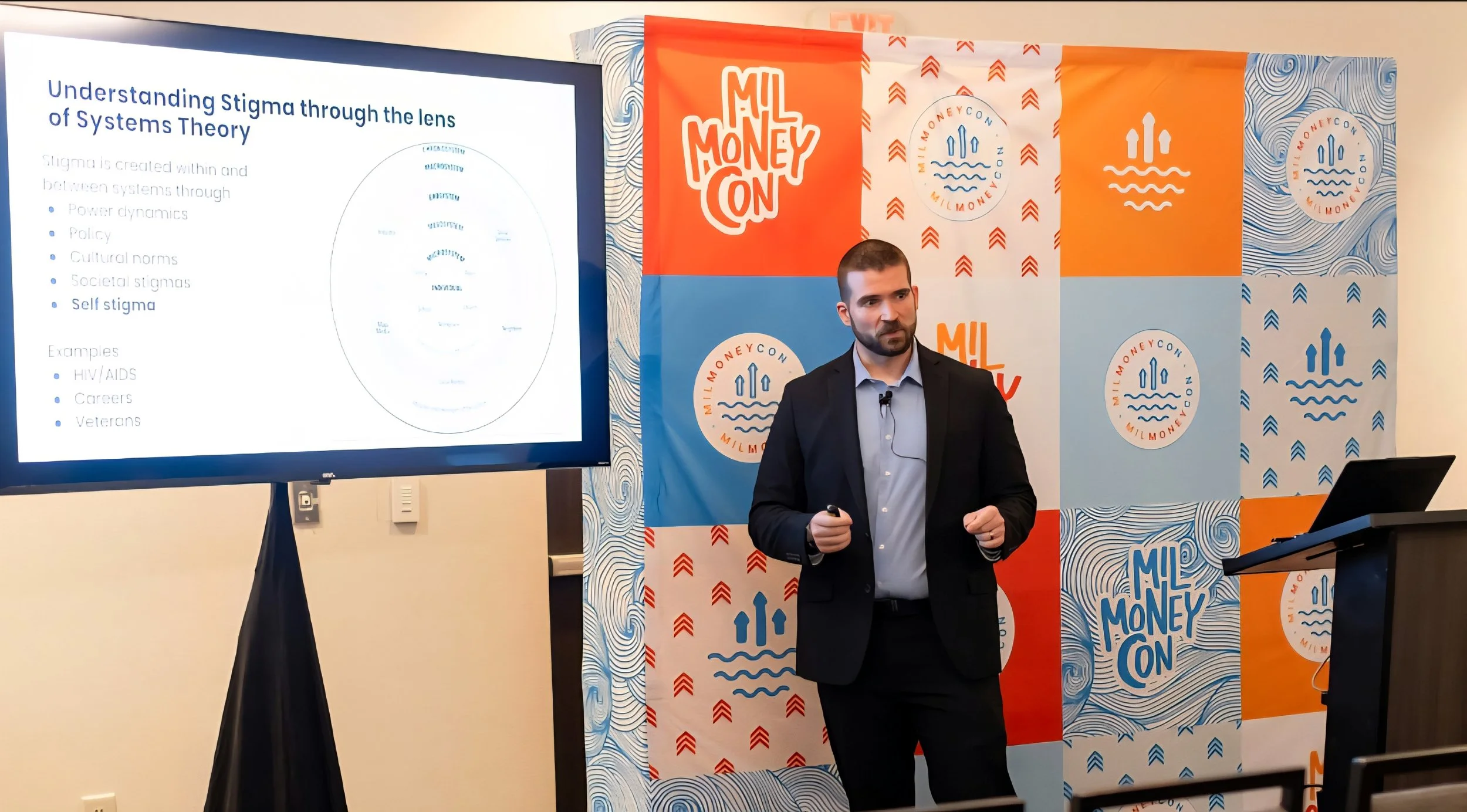 A man in a black suit giving a presentation at a conference with a slide about stigma and systems theory on a large screen, and a colorful background with logos reading "ML Money on".