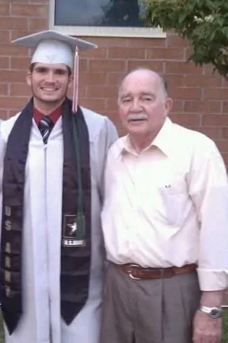 A young man in a graduation cap and gown standing next to an older man in a light-colored shirt in front of a brick building and tree.
