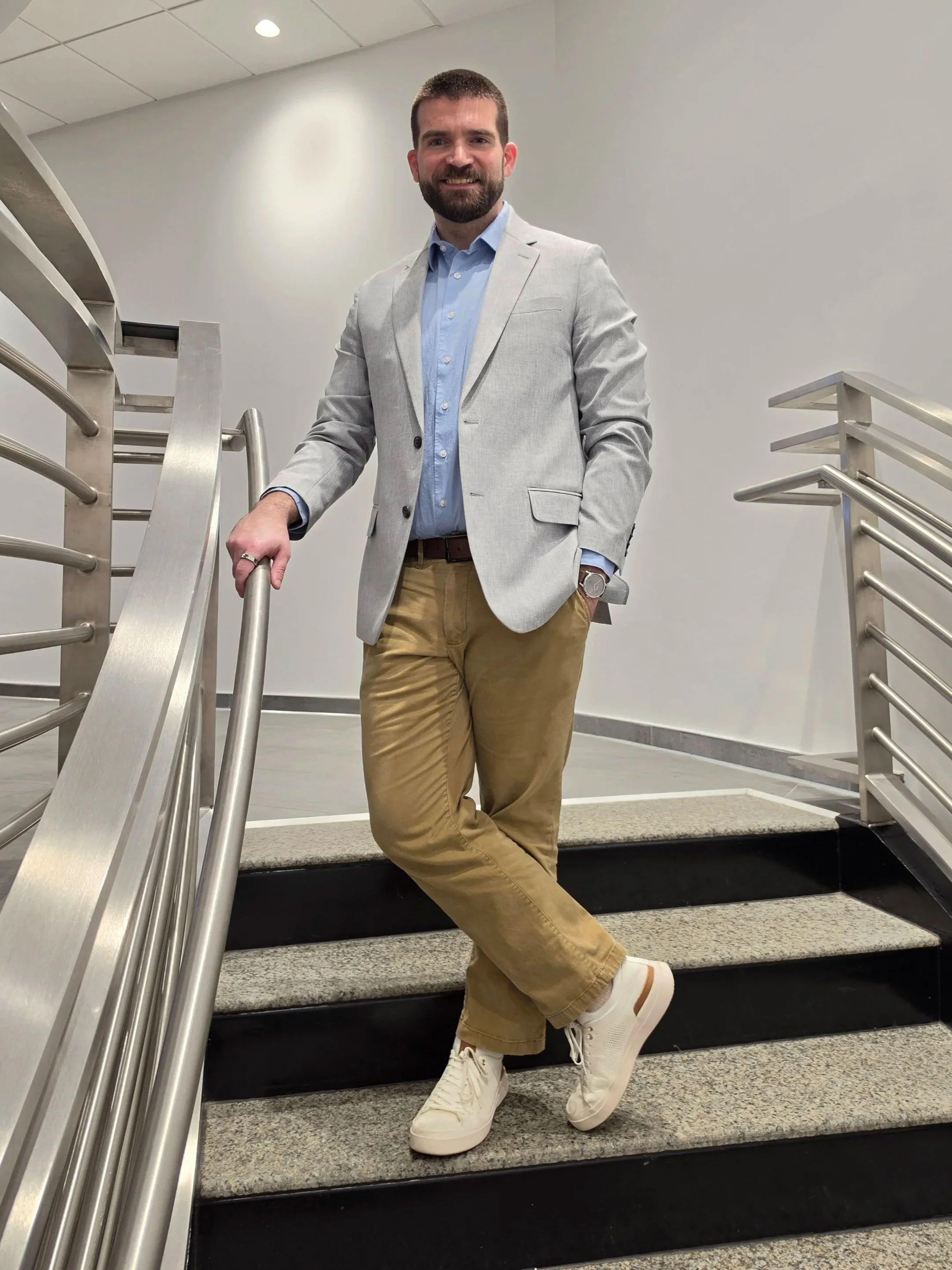 A man with a beard, wearing a light gray blazer, light blue shirt, khaki pants, white sneakers, and a watch, posing on stairs in an indoor setting with metal handrails and a gray wall.