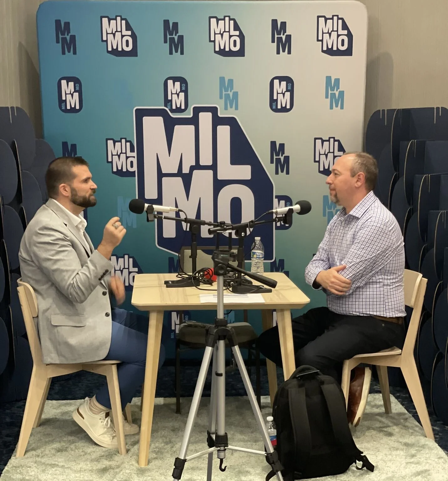 Two men are seated at a small table in a podcast studio, engaged in an interview. The man on the left, wearing a light-colored blazer, blue pants, and white sneakers, is speaking into a microphone. The man on the right, dressed in a checkered shirt a