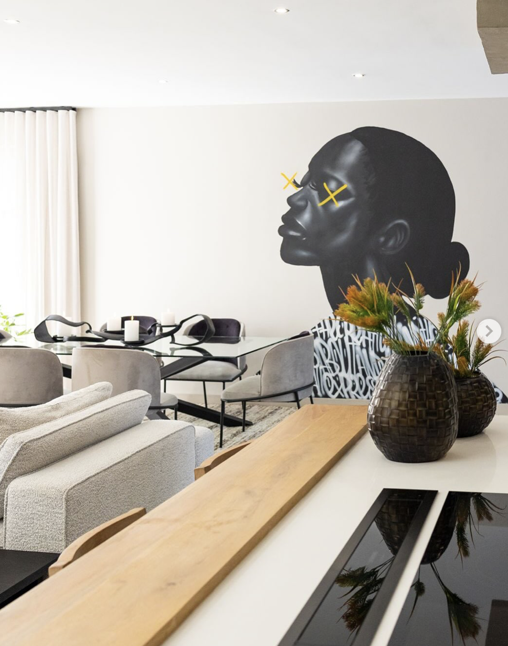 Modern living and dining room with a large black and white mural of a woman's profile on the wall, yellow X's over her eyes, a white table, grey chairs, beige sofa, candle holders, a wooden tray on a white surface, and two dark baskets with orange and green flowers.