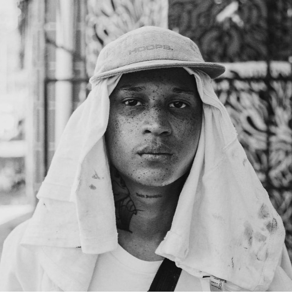 A black and white portrait of a person wearing a cap and a cloth draped over their head and shoulders, with tattoos and freckles visible on their face, standing outdoors in front of a textured wall.