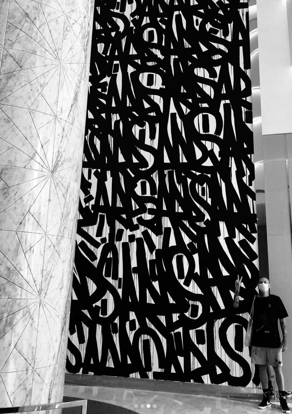 A person standing in front of a large black and white abstract wall art with bold, overlapping letters, inside a modern building with marble wall on the left.