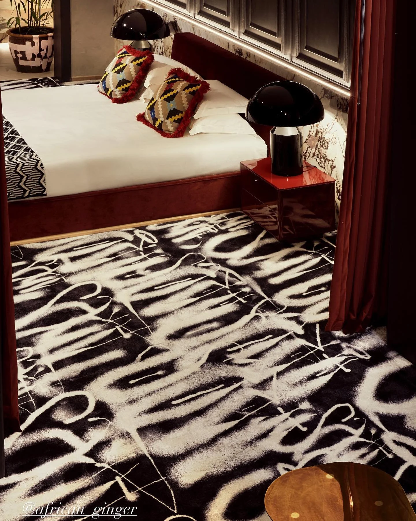 A bedroom with a bold black and white animal print rug, a bed with white bedding and patterned pillows, dark red curtains, a dark headboard, unique black bedside lamps on red nightstands, a plant in a decorative pot, and a small wooden table.