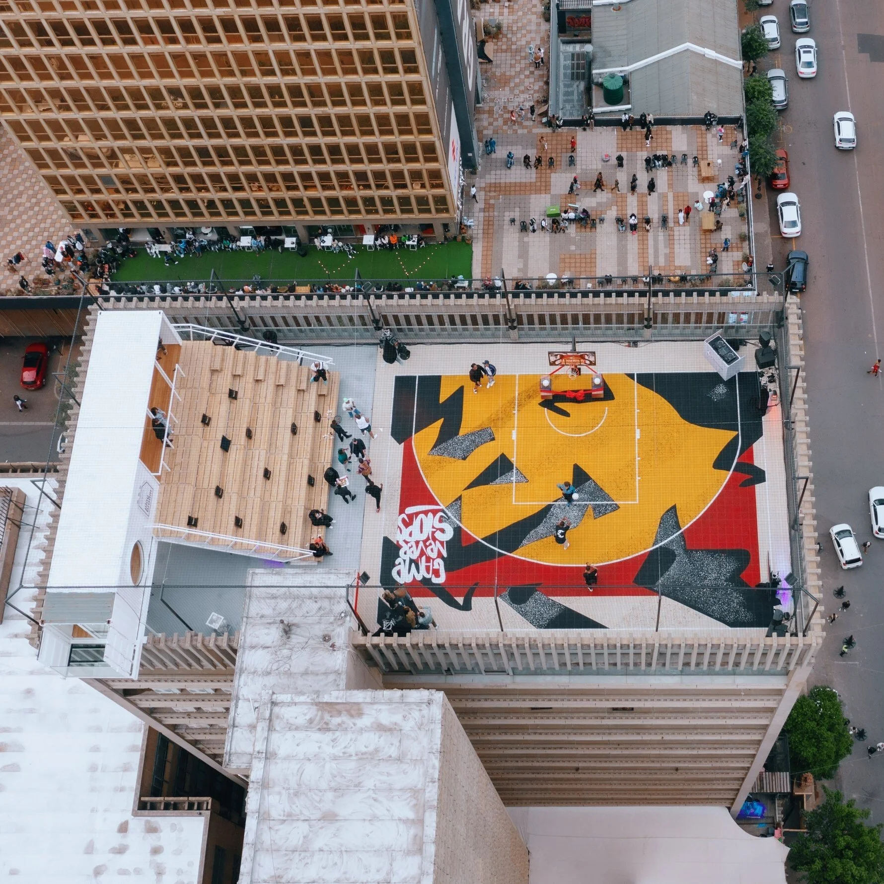 An aerial view of an urban rooftop with a basketball court featuring a colorful design, people socializing and walking on a patio, and a parking lot with cars. The scene includes a modern building with wooden framing and a smaller adjacent structure.