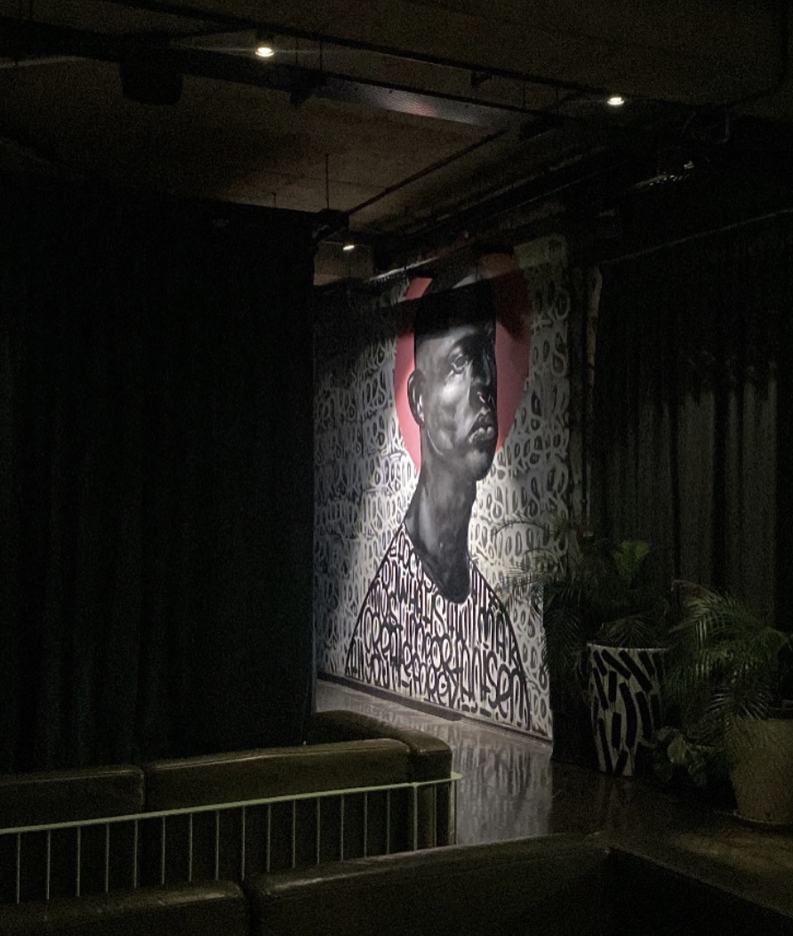 A mural of a black man with short hair, painted in grayscale, is displayed on a wall with a pink background and black and white patterned background behind. The mural is on a stage or wall with black curtains and indoor plants nearby.