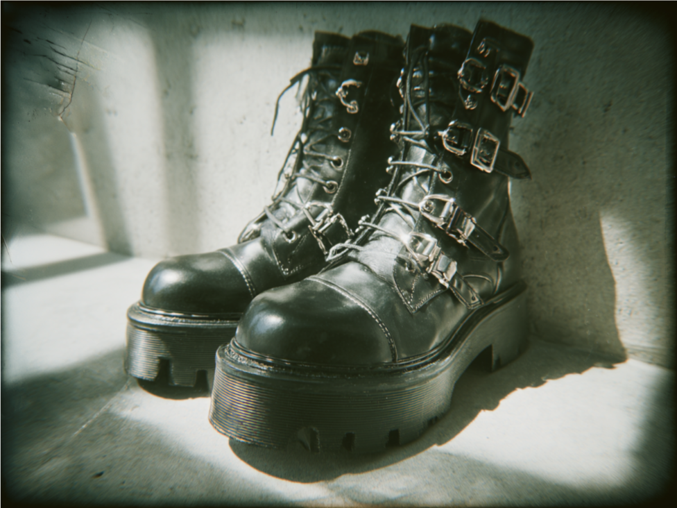 Pair of black leather combat boots with buckles and thick soles, placed on a concrete floor near a wall with sunlight casting shadows.