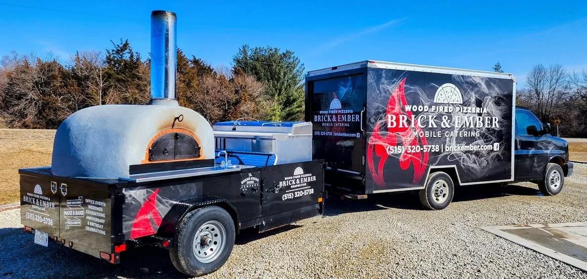 Food truck with a wood-fired pizza oven on a trailer, parked outdoors on gravel with trees in the background, advertising Brick & Ember mobile catering.