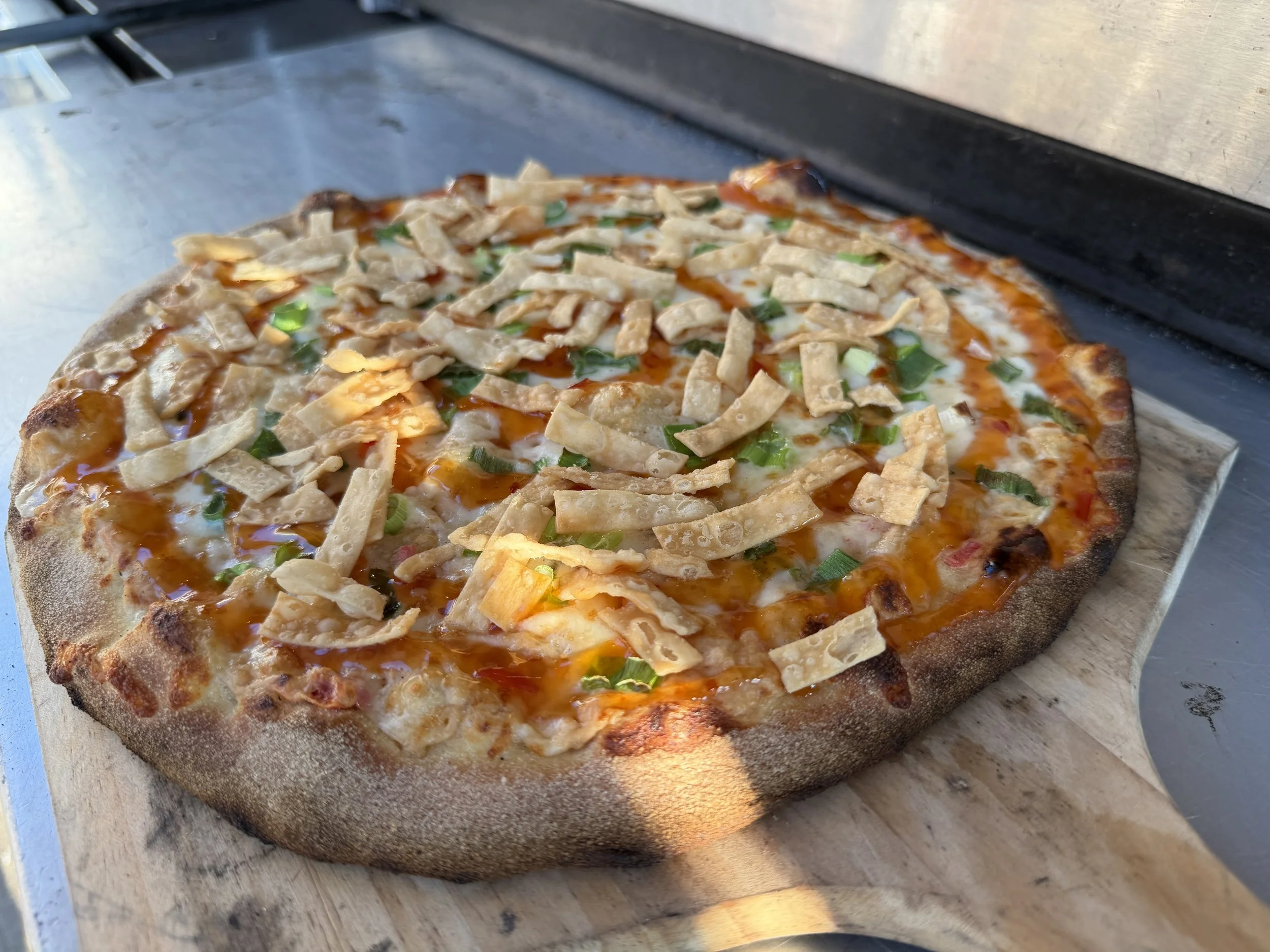 Close-up of a freshly baked pizza with chicken, green onions, cheese, and hot sauce, resting on a wooden pizza peel.