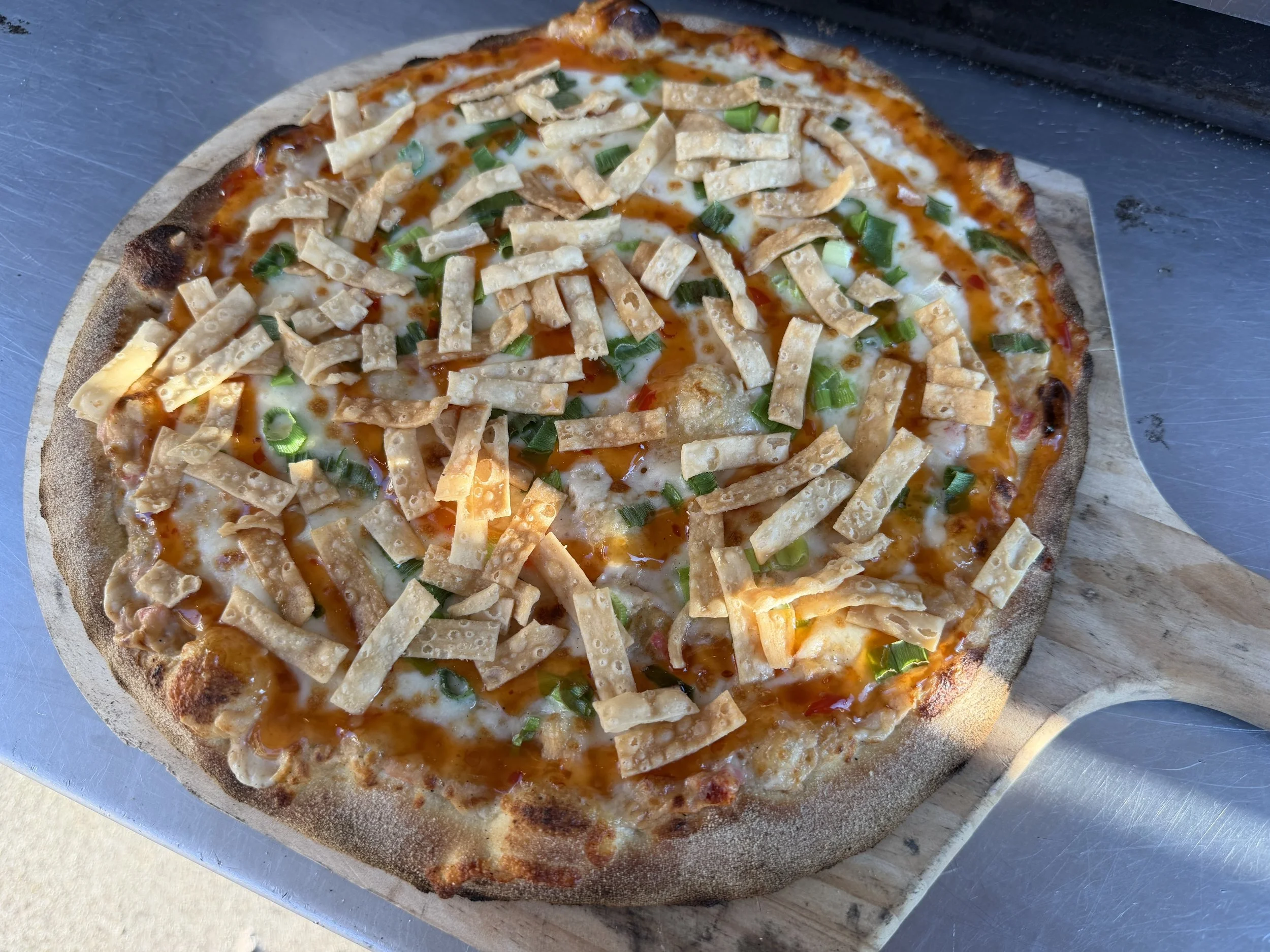 A pizza with cheese, chicken strips, green onions, and a drizzle of hot sauce on top.
