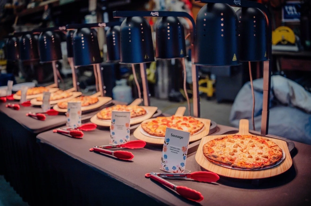 A row of pizzas on wooden boards under heat lamps at a pizza buffet station, with red tongs placed beside each pizza and small signs labeling the pizza toppings, such as pepperoni and sausage.