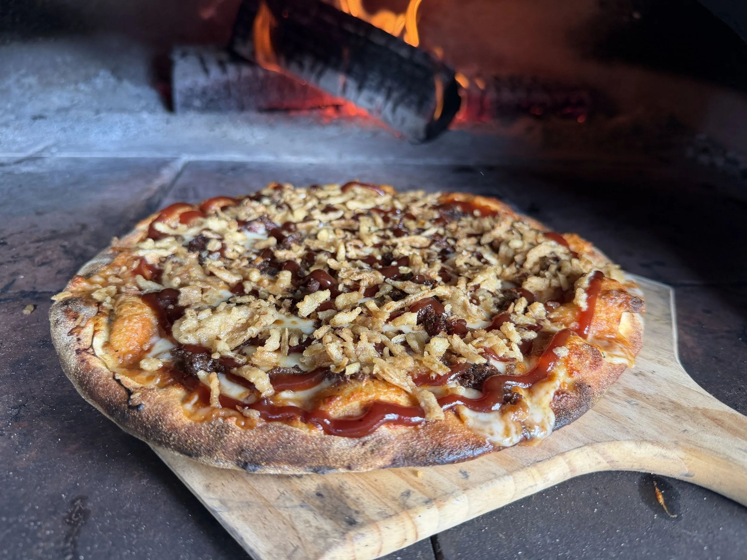 Pizza with toppings of cheese, chicken, beans, and barbecue sauce on a wooden board inside an oven.